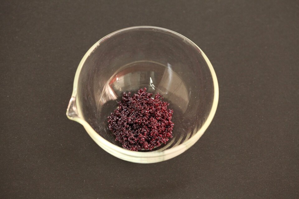 Na zdjęciu widoczne jest szklane okrągłe naczynie, na dnie którego umieszczona jest brunatnoczerwona, grudkowata substancja.