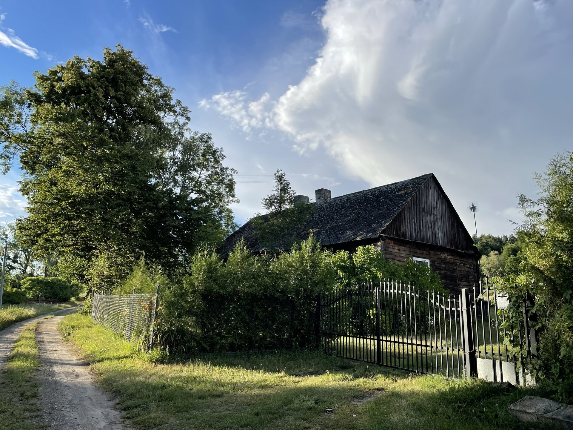 Fotografia przedstawia drewnianą chatkę z drewnianym płotem. Brama pomalowana na ciemnożółty kolor, a pośrodku dla dekoracji namalowano na niej dwa brązowe romby z białymi konturami.