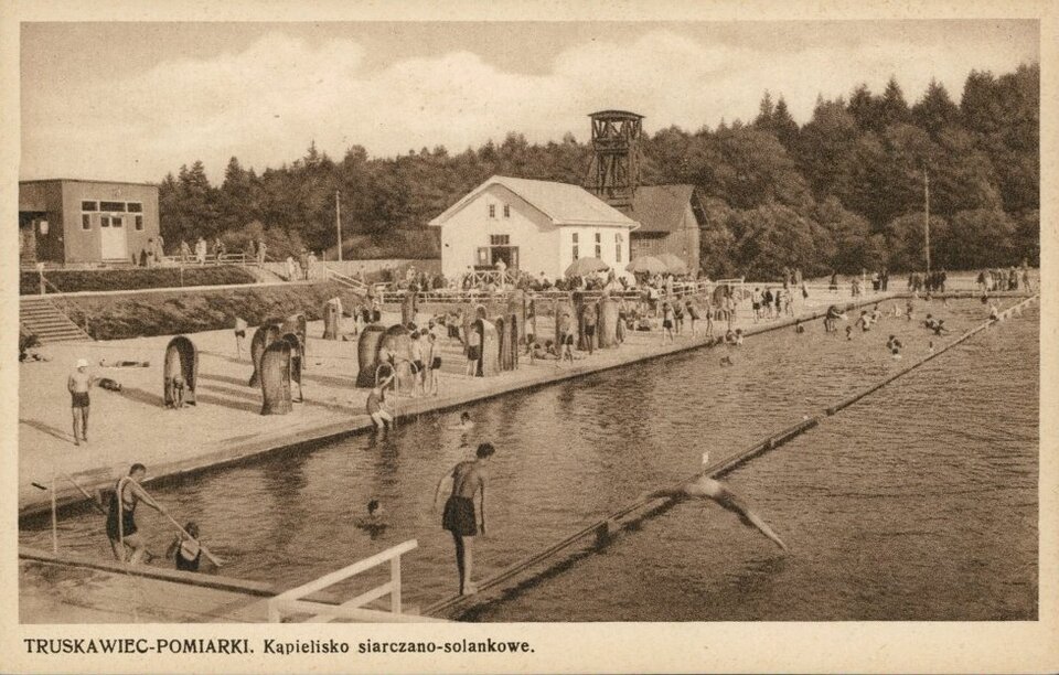 Zdjęcie przedstawia basen na świeżym powietrzu. Pływają w nim ludzie. Nad brzegiem jest plaża z siedziskami. W tle jest las.