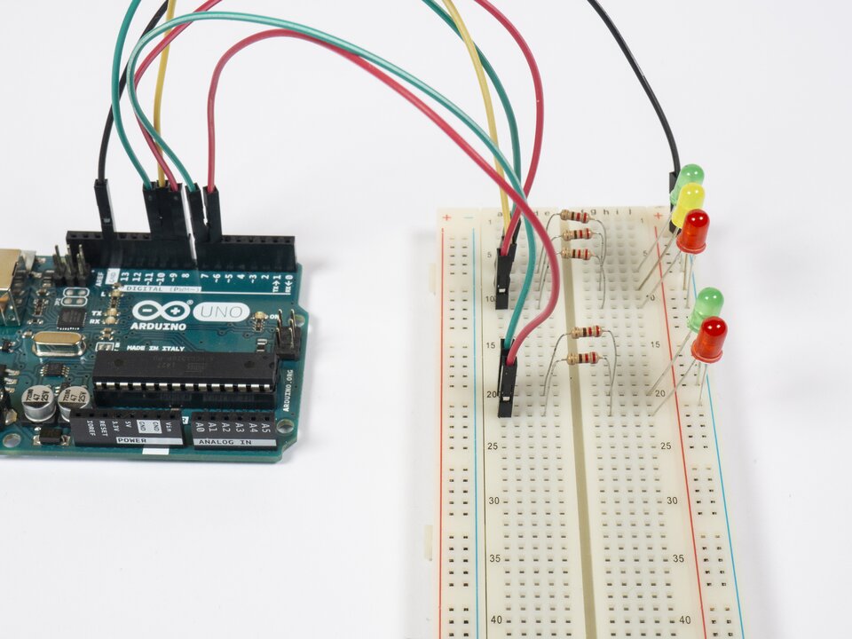 Zdjęcie przedstawia moduł Arduino z płytką stykową połączone podpiętymi przewodami. Na zdjęciu znajdują się pięć wpiętych rezystorów 220 Ω omega oraz wpięte do płytki stykowej diody LED: zielona, żółta i czerwona symulujące światła dla pojazdów, następnie dalej zielona i czerwona symulujące światła dla pieszych. Przewodami połączono piny z płytką stykową.