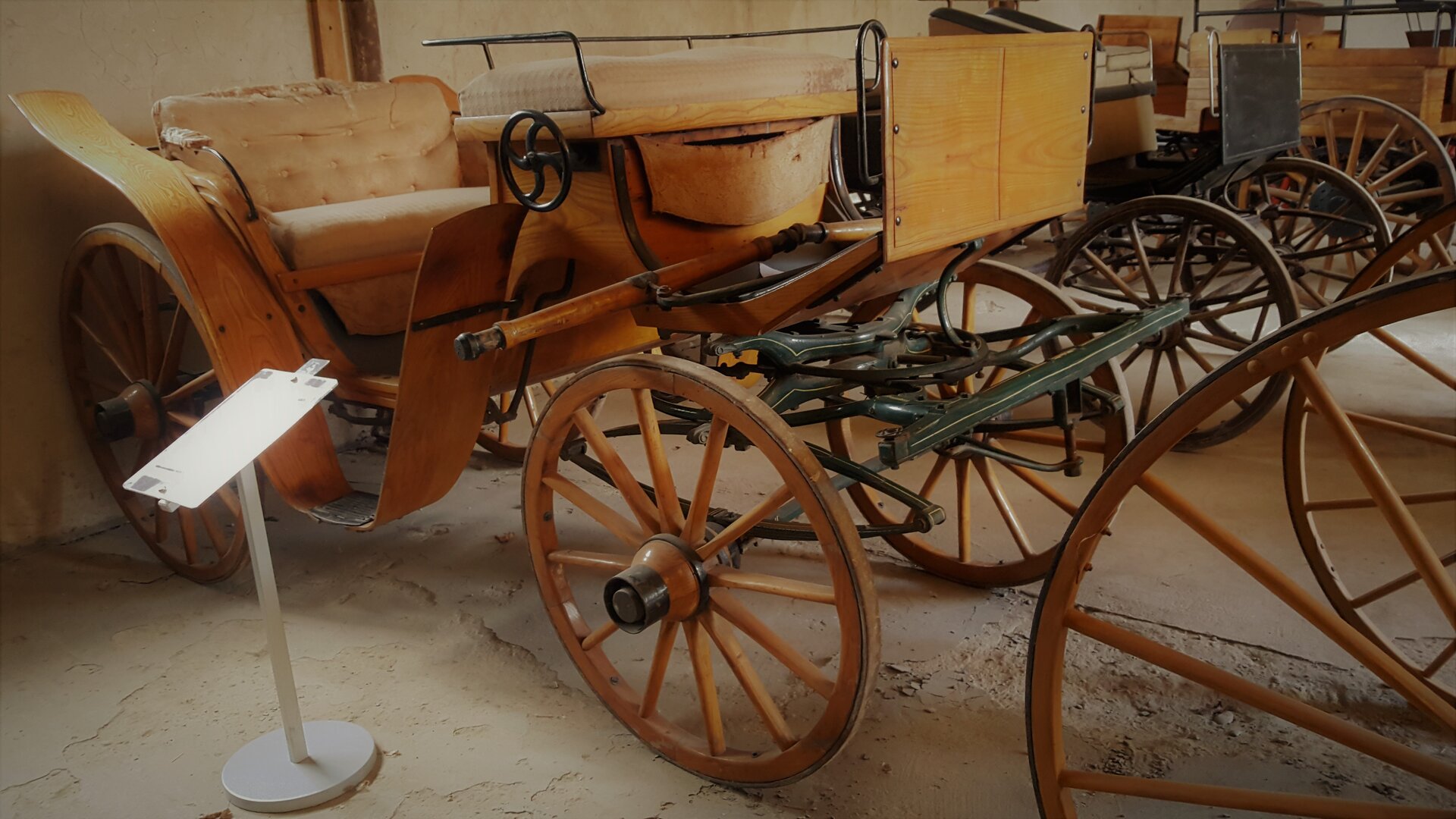 Zdjęcie przedstawia zabytkowy wolant stojący na wystawie. Jest to czterokołowy, drewniany, lekki powóz bez zadaszenia. 
Szczegółowy opis uzupełniający zdjęcie znajduje się w treści definicji.