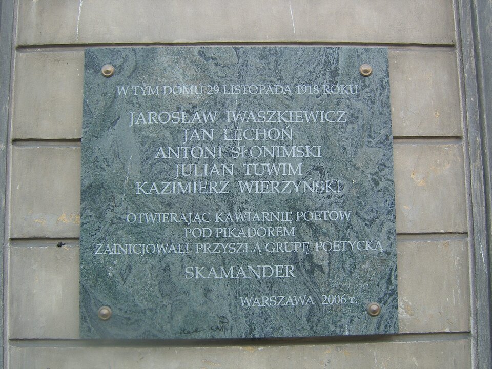 Fotografia przedstawia tablicę pamiątkową wiszącą na ścianie budynku. Umieszczono na niej napis: W tym domu 29 listopada 1918 roku Jarosław Iwaszkiewicz, Jan Lechoń, Antoni Słonimski, Julian Tuwim, Kazimierz Wierzyński, otwierając kawiarnię poetów Pod Pikadorem zainicjowali, przyszłą grupę poetycką Skamander. Warszawa 2006 roku.