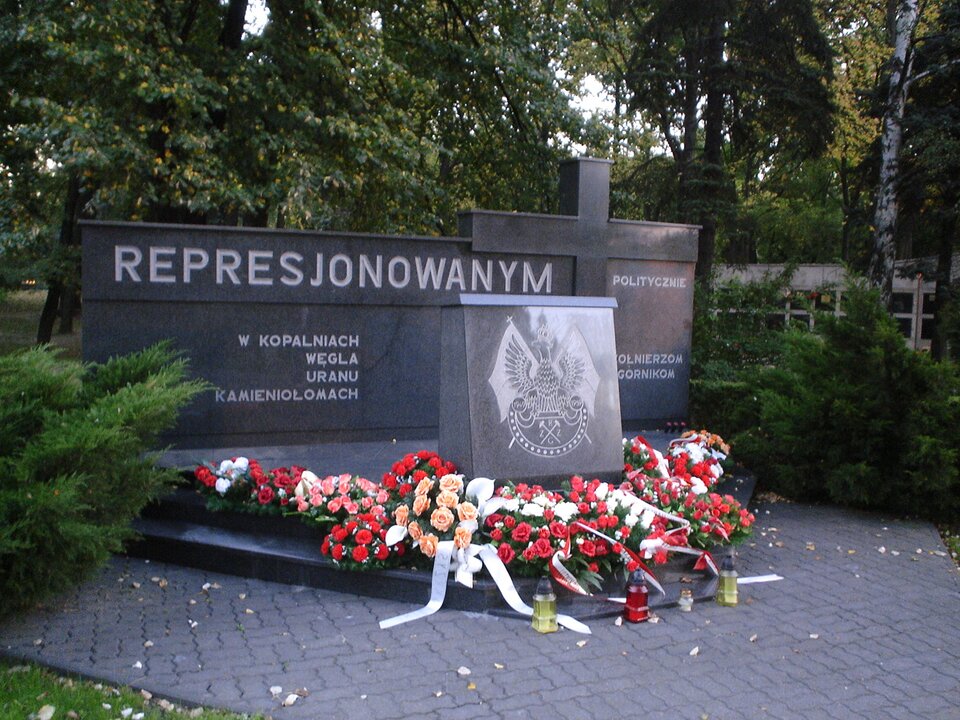 Ilustracja przedstawia pomnik z czarnego materiału. Na pionowej płycie napis: Represjonowanym politycznie. W kopalniach węgla, uranu, kamieniołomach - żołnierzom, górnikom. Obok krzyż. Z przodu tabliczka z orłem w koronie. Pod nią wiązanki kwiatów. 