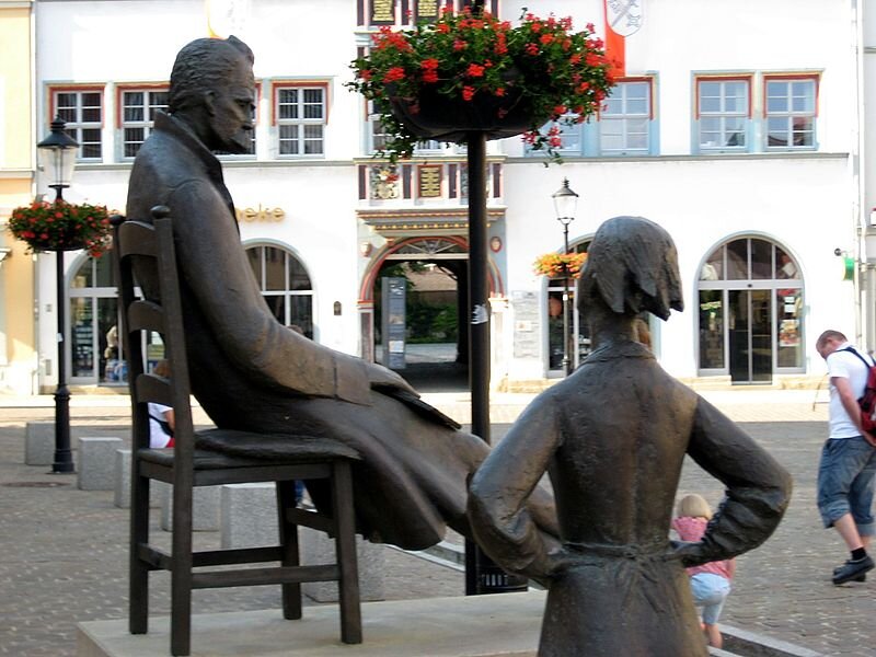 Zdjęcie przedstawia rzeźbę mężczyzny w sile wieku siedzącego na krześle. Postać pochyla głowę, ukazuje lewy profil pociągłej twarzy. Mężczyzna ma proste, zaczesane ku górze włosy. Ma wysokie, lekko zmarszczone czoło i gęste brwi. Jego oczy są głęboko osadzone. Ma niewielki nos. Na twarzy widać gęste, długie wąsy. Ubrany jest w długi płaszcz. Ręce opiera o kolana. Ma wyprostowane nogi. Na pierwszym planie ukazana jest rzeźba przedstawiająca kobietę opierającą dłonie o biodra. Kobieta stoi tyłem do widza. Ma krótkie włosy. W głębi, za rzeźbami stoi trzykondygnacyjna kamienica. Na parterze są duże, łukowate witryny i brama pośrodku. Okna na pierwszym piętrze są niewielkie. Budynek jest oświetlony.