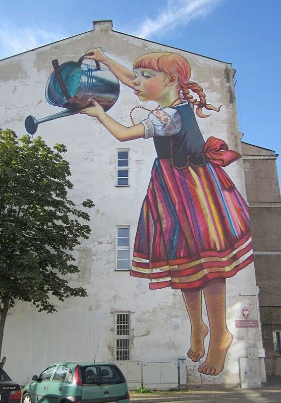 Fotografia przedstawia mural na jednym z budynków w Białymstoku: dziewczynkę trzymającą w dłoniach konewkę. Obok ściany budynku z muralem znajduje się drzewo. Tworzy to wrażenie, że dziewczynka z muralu podlewa prawdziwe drzewo. 