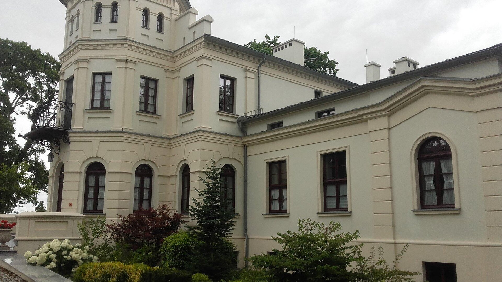 Zdjęcie przedstawia żółty dworek. Budynek z prawej strony jest parterowy, a z lewej dwupiętrowy. Jego przód jest zwieńczony dwupiętrową wieżą. Przed budynkiem rosną zielone krzewy. Z prawej strony zdjęcia widoczny jest fragment drzewa. Niebo jest zachmurzone. Zdjęcie zostało wykonane w świetle dziennym.