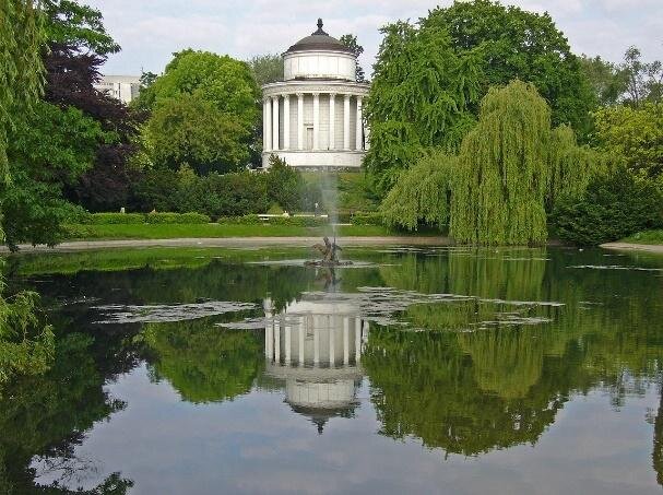 Zdjęcie przedstawia niewielkie jeziorko, na brzegu którego znajduje się niewielka fontanna. Za nią znajduje się niewielki, okrągły, biały budynek. Jest on ozdobiony kolumnami. Wokół niego rosną drzewa liściaste. 
