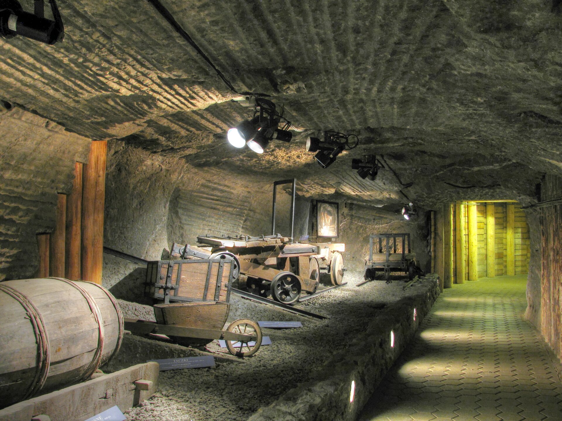 Zdjęcie ukazuje wystawę na trasie turystycznej. Są tam między innymi: drewniana beczka, drewniana taczka, wagoniki kopalniane.