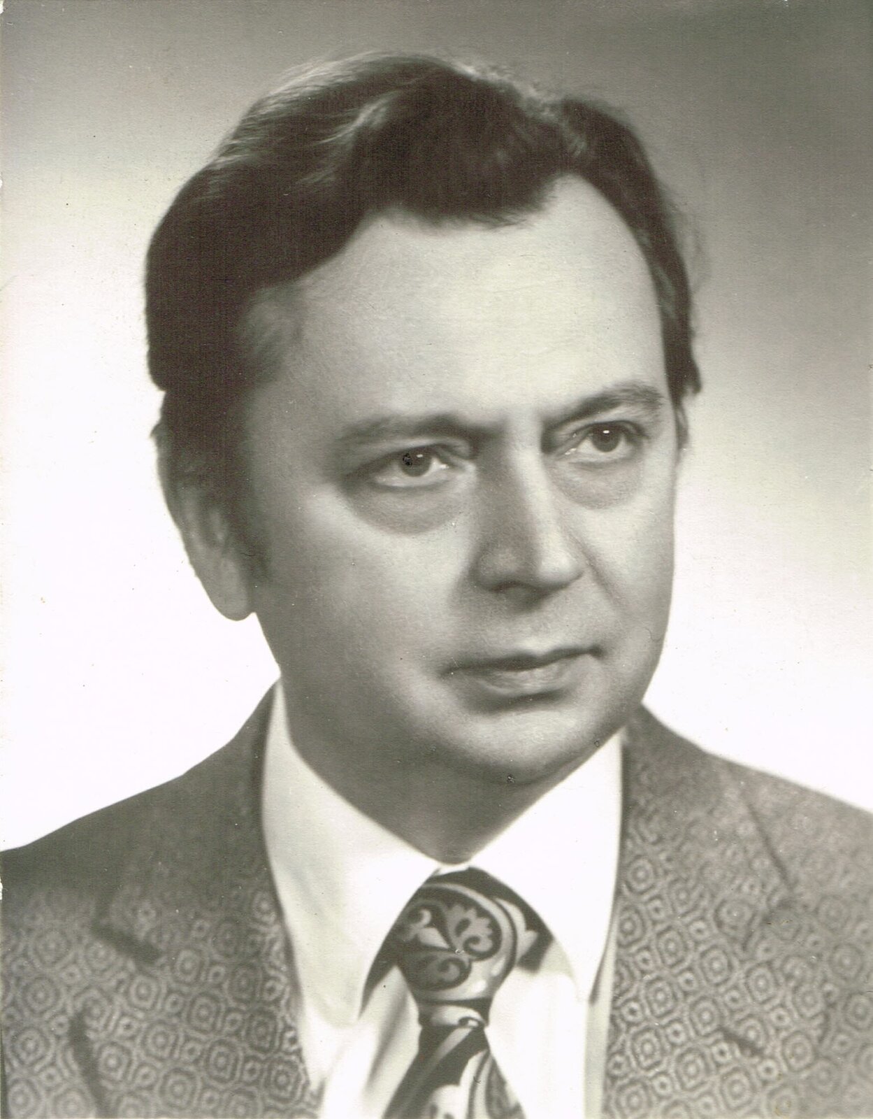 Ilustracja przedstawia czarno‑białą fotografię Karola Musioła. Mężczyzna z poważną miną w  średnim wieku o czarnych włosach, wysokim czole oraz podkrążonymi oczami. Ubrany jest w marynarkę z wzorami w kształcie rombów, białą koszule oraz elegancki czarny krawat z jasnymi wzorami. 