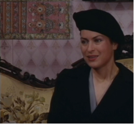 Zdjęcie przedstawia kobietę w czarnym stroju z czarnym beretem na głowie. Siedzi na wzorzystej kanapie i mówi.