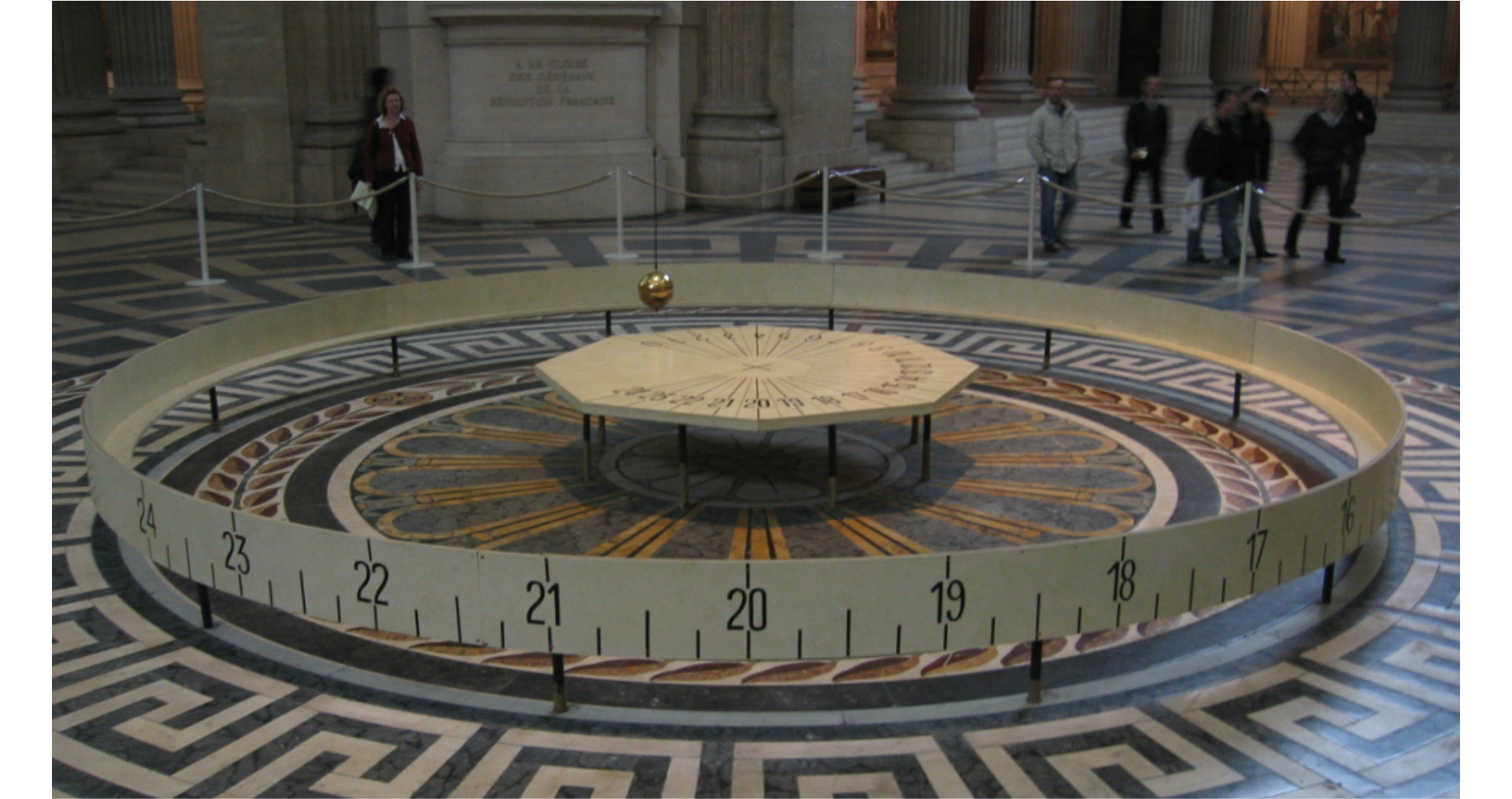 Zdjęcie przedstawia wnętrze wysokiego budynku. We wnętrzu budynku znajduje się ogrodzona strefa, wewnątrz której, nad ośmiokątnym stołem, waha się wahadło Foucaulta. Na stole, wokół jego osi, narysowana jest podziałka. Ponieważ strefa jest ogrodzona i zwiedzający nie mają możliwości odczytania liczb z podziałki na stole, została ona skopiowana na stojącą wokół stołu wstęgę.