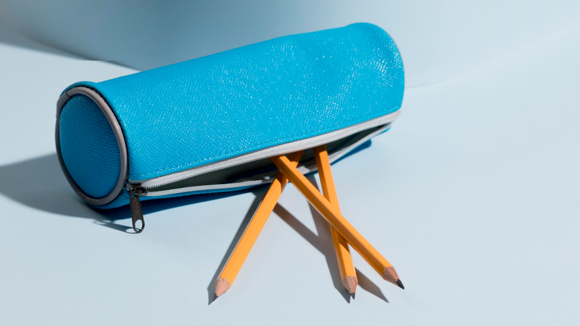 Zdjęcie przedstawia materiałowy piórnik w kształcie podłużnego walca. Ekspres piórnika jest rozsunięty. Z piórnika wystają trzy takie same ołówki.