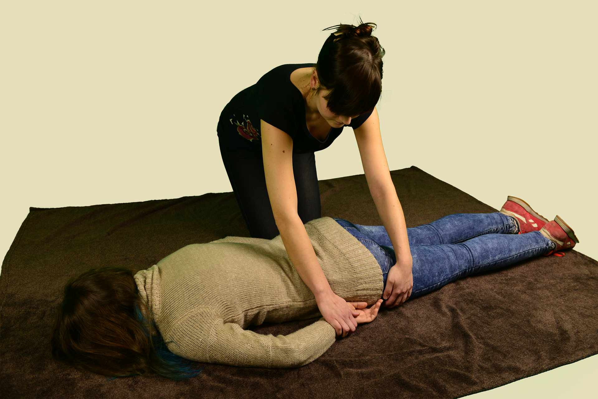 Poszkodowana osoba leży na brzuchu. Ręce ułożone wzdłuż tułowia. Nogi proste, równolegle do siebie. Twarz zwrócona do podłoża. Osoba leży zwrócona głową w lewą stronę zdjęcia. Obok poszkodowanej osoby, w głębi zdjęcia, klęczy kobieta układająca poszkodowaną osobę w pozycji bezpiecznej. Kobieta przytrzymuje dłoń i biodro osoby leżącej. Dłoń wkłada pod biodro.