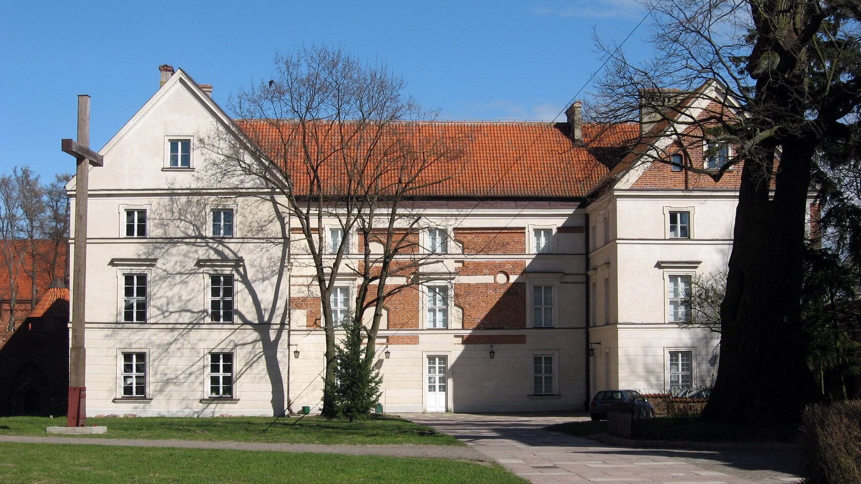 Zdjęcie przedstawia dwupiętrowy budynek. Budynek jest w kształcie litery C. Dach ma dwuspadzisty, przykryty czerwoną dachówką. W środkowej części elewacji widać cegły – na wysokości drugiego i trzeciego piętra. Przed budynkiem, po lewej stronie stoi wysoki krzyż. Widoczne są trzy stare drzewa. Dwa z nich umiejscowione są bliżej budynku, w środkowej części. Pozostałe jest po prawej stronie, przed budynkiem. Do budynku prowadzi szeroki chodnik. 