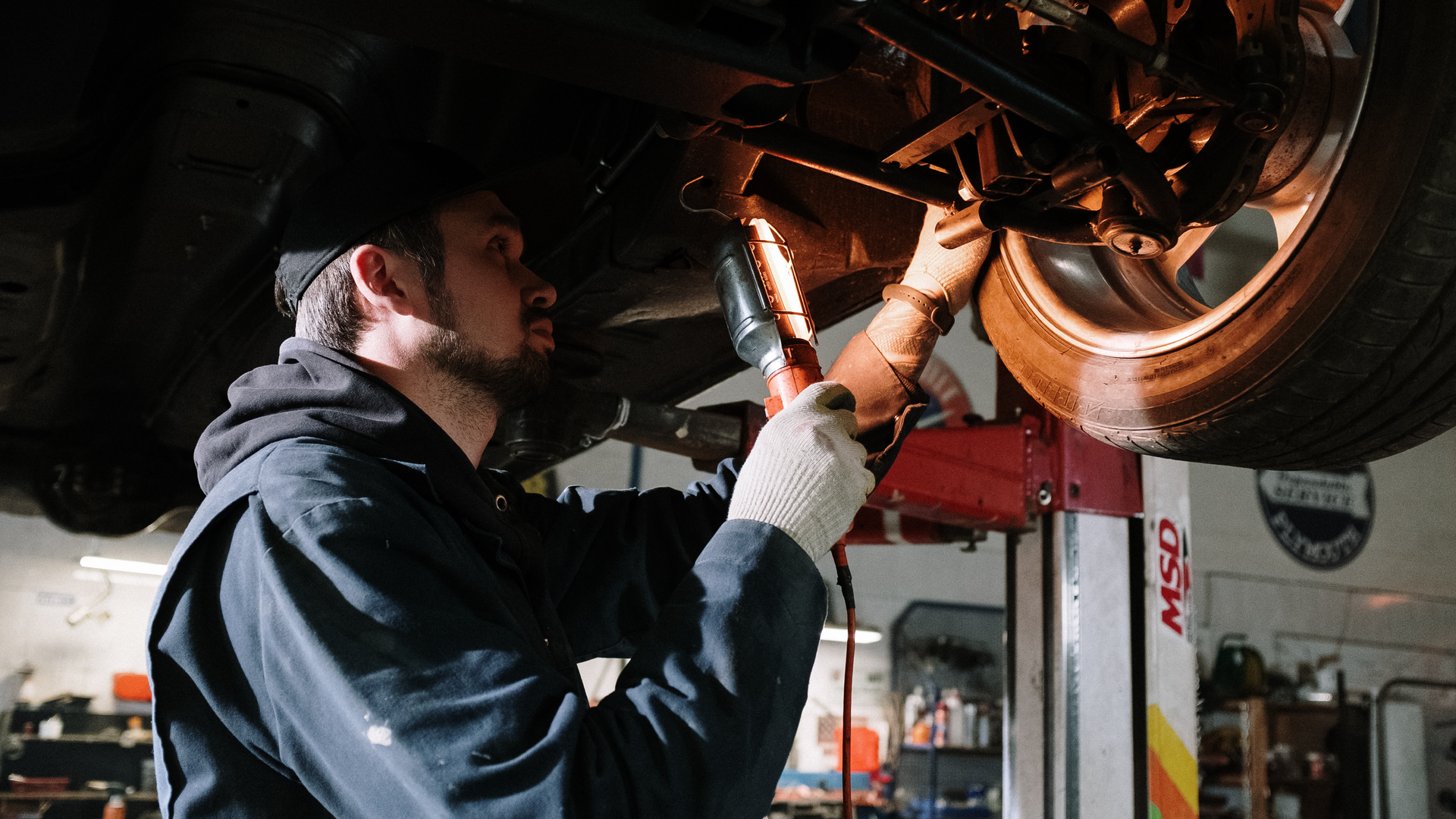 Zdjęcie przedstawia młodego mężczyznę z brodą. Mężczyzna znajduje się pod samochodem na podnośniku i oświetla lampą podwozie pojazdu.