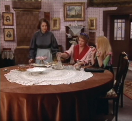 Zdjęcie przedstawia trzy postaci znajdujące się przy drewnianym stole. Kobieta z ciemnymi włosami i ciemnoniebieską koszulą i jasnym fartuchu stoi. Pozostałe kobiety siedzą. Jedna ma różową podomkę, a druga blond włosy i zieloną bluzkę. Na stole jest haftowany obrus.