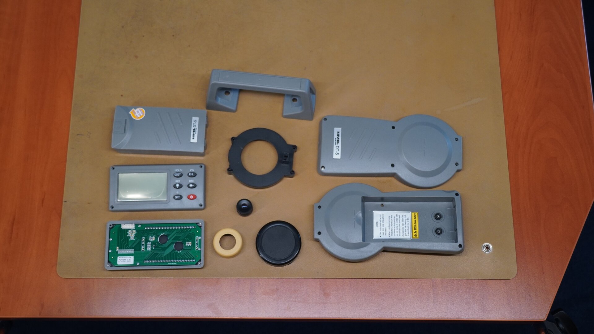 Na zdjęciu pokazano zdemontowane części teodolitu: baterię, panel sterujący z wyświetlaczem, płytkę modułu sterującego, górny uchwyt urządzenia, pierścień obiektywu, pierścień okularu, przysłonę obiektywu, boczną osłonę, drugą boczną osłonę z gniazdem na baterię.