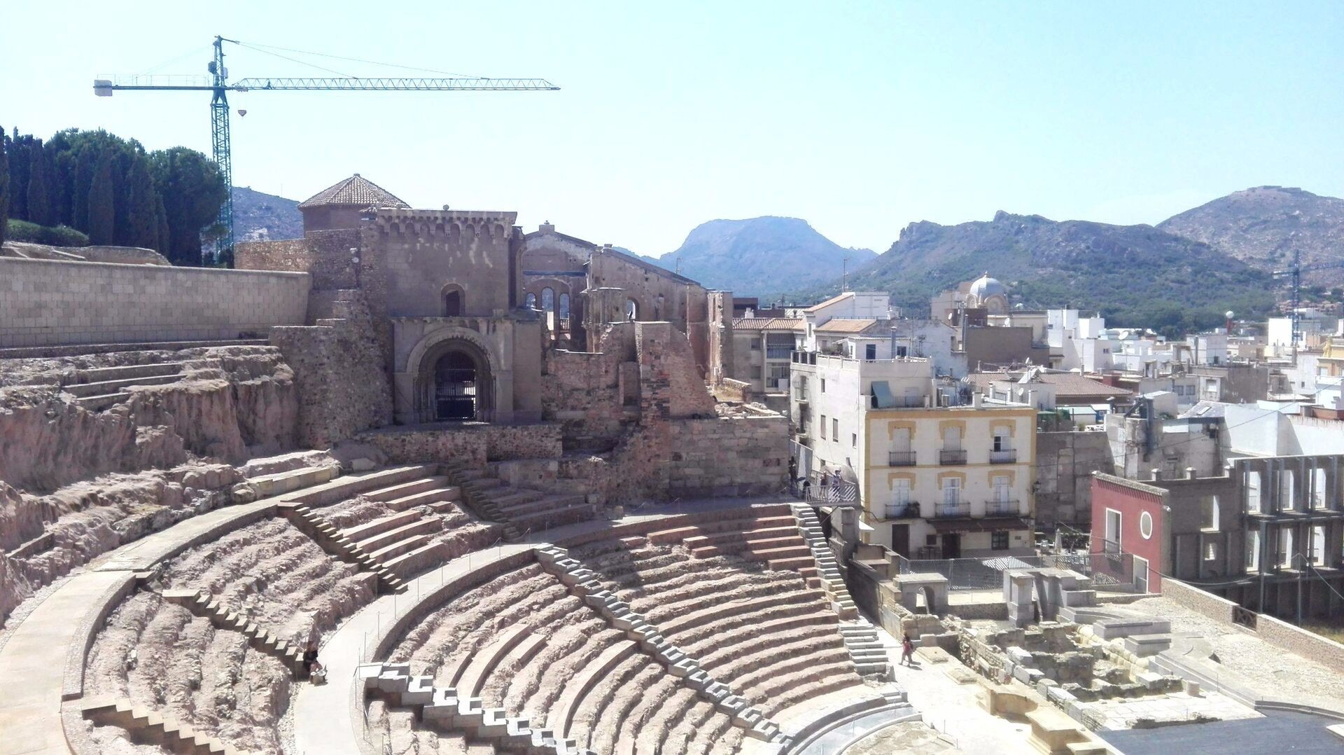 Ilustracja przedstawia teatr rzymski w Kartaginie znajdujący się w centrum miasta. Teatr ma półkolisty kształt. Widownia została podzielona poziomymi przejściami oraz biegnącymi promieniście schodkami na sektory poziome i pionowe. Obiekt otoczony jest współczesną zabudową. W tle widoczny jest dźwig.