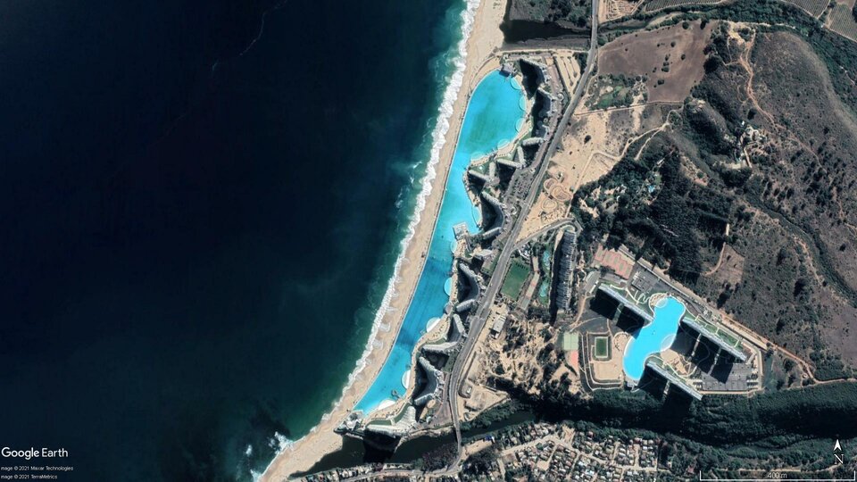 Zdjęcie przedstawia widok satelitarny na kurort. Z lewej strony jest morze, następnie wąski pas piasku. Za nim jest basen, nad którym wznoszą się zabudowania kurortu. Wyżej biegnie droga, za którą jest dalsza część kurortu z mniejszym basenem. Wokół kurortu są tereny porośnięte zielenią.