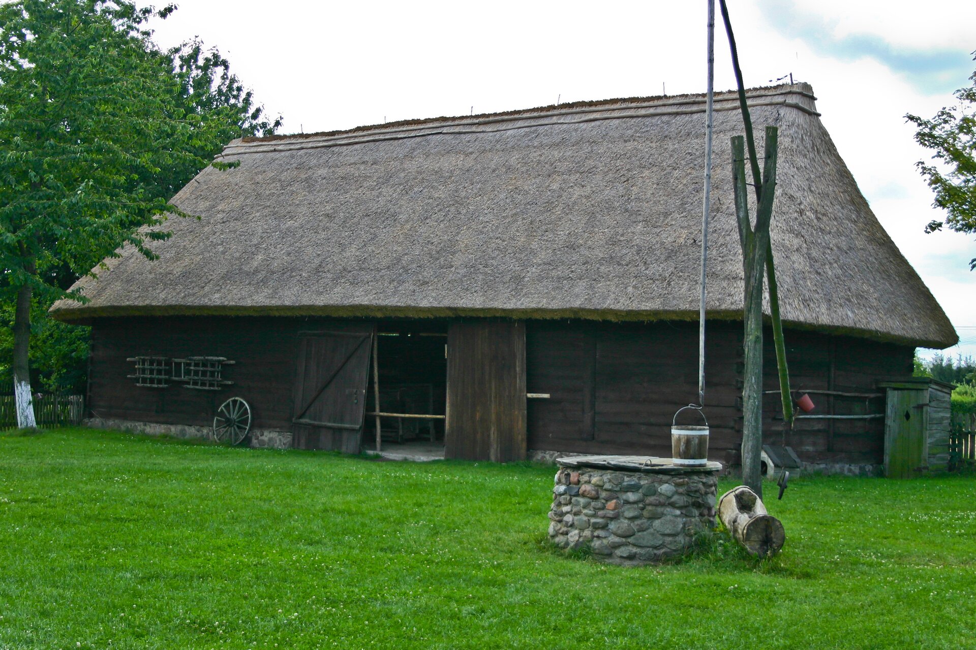 Fotografia przedstawia stodołę należącą do Wielkopolskiego Parku Etnograficznego w Dziekanowicach. Ukazuje drewniany obiekt kryty strzechą. Przed stodołą stoi żuraw z kamienną studnią. Dookoła rośnie trawa. Obok stodoły stoi drewniany wychodek. Po lewej stronie obiektu rośnie duże drzewo.