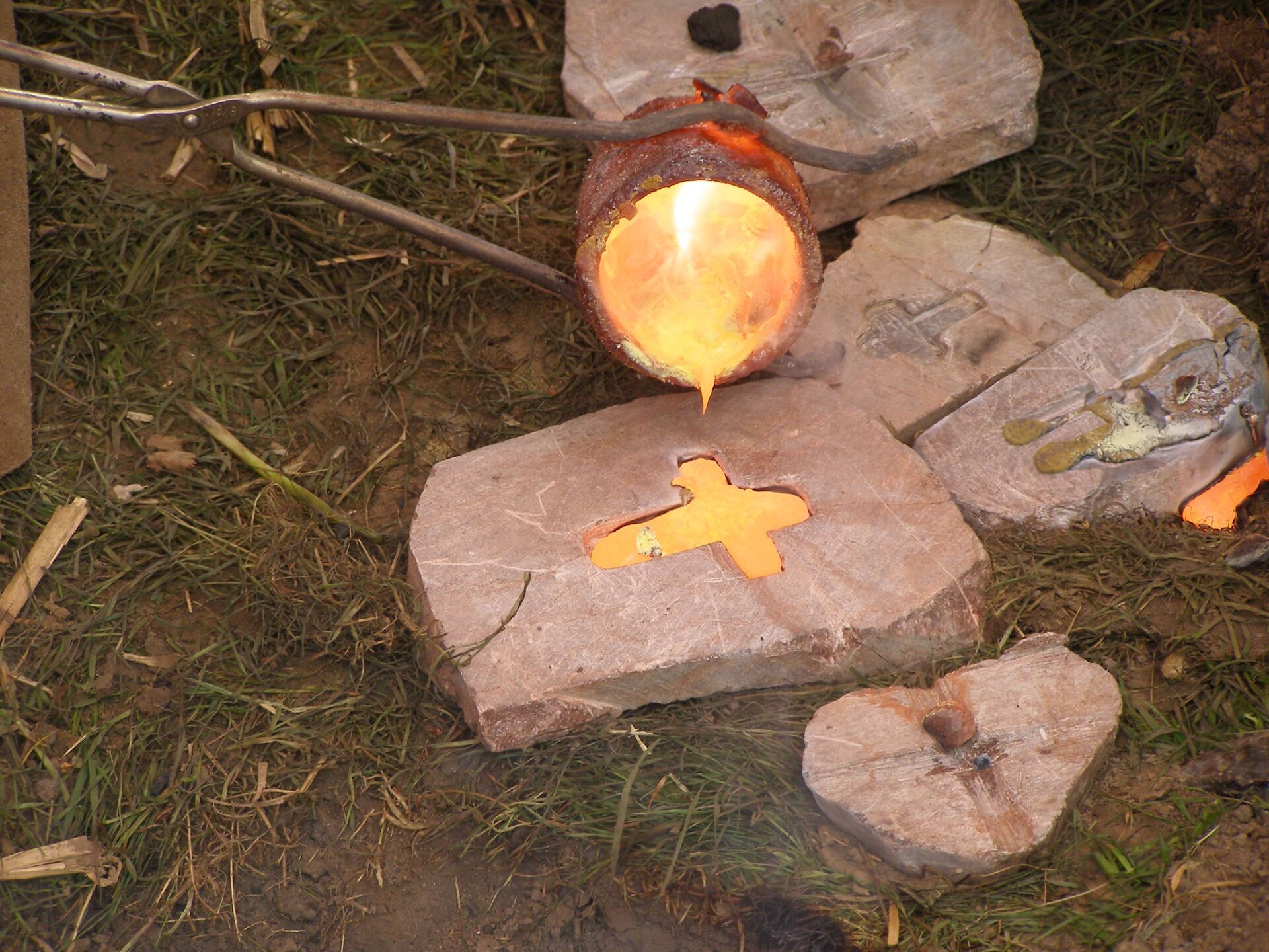 Zdjęcie przedstawia odlewanie przedmiotu z brązu dawnymi metodami. Na obrośniętej trawą ziemi leży wycięta w kamieniu forma w kształcie krzyżyka, do której z trzymanego w długich szczypcach małego tygla nalewany jest płynny metal. Zarówno zawartość formy, jak i tygla jarzą się pomarańczowo‑żółtym blaskiem. Wokół na trawie porozrzucane są inne, popsute formy.
