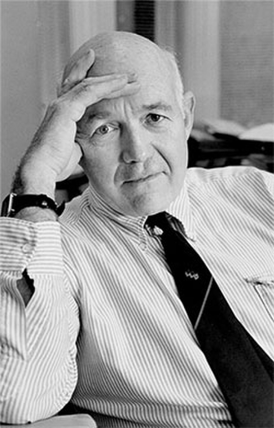 Ilustracja przedstawia mężczyznę w starszym wieku. Siedzi on przy stole podpierając głowę dłonią. Ubrany jest w koszulę i krawat. Ma pogodną, zamyśloną twarz, duże oczy, duże brwi, łysinę. Mężczyzna spogląda do przodu.