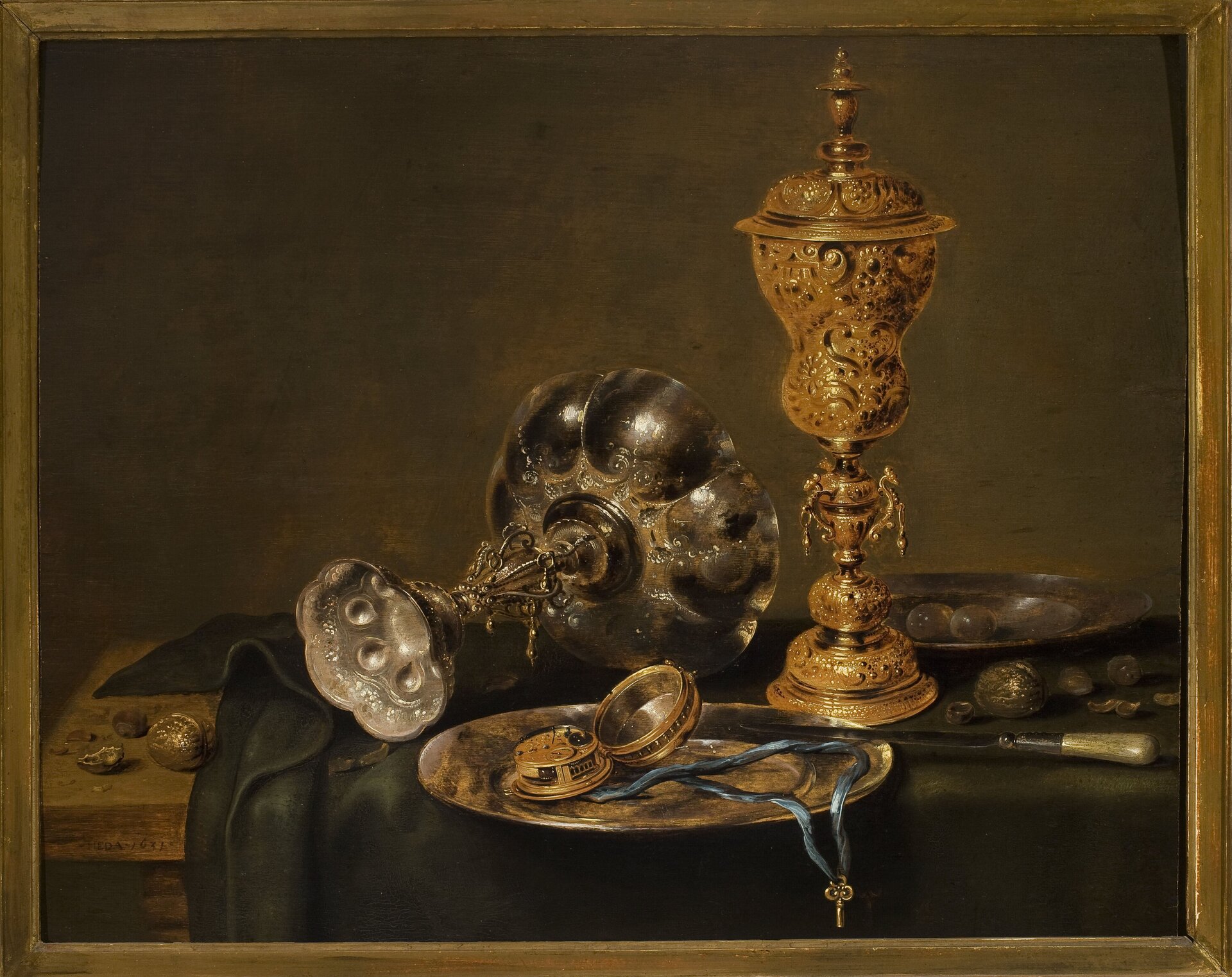 Obraz przedstawia kuchenne naczynia. Na stole znajduje się ciemnozielony obrus, na którym stoi talerz. Na nim znajduje się błękitny sznurek na którego końcu zawieszony został medalik w kolorze złotym. O krawędź talerza oparty został nóż z białą rękojeścią. Następnie po prawej stronie talerza stoi bogato zdobiony puchar. Jest on wykonany ze złota. Po lewej stronie stołu w pozycji leżącej znalazł się srebrny puchar.