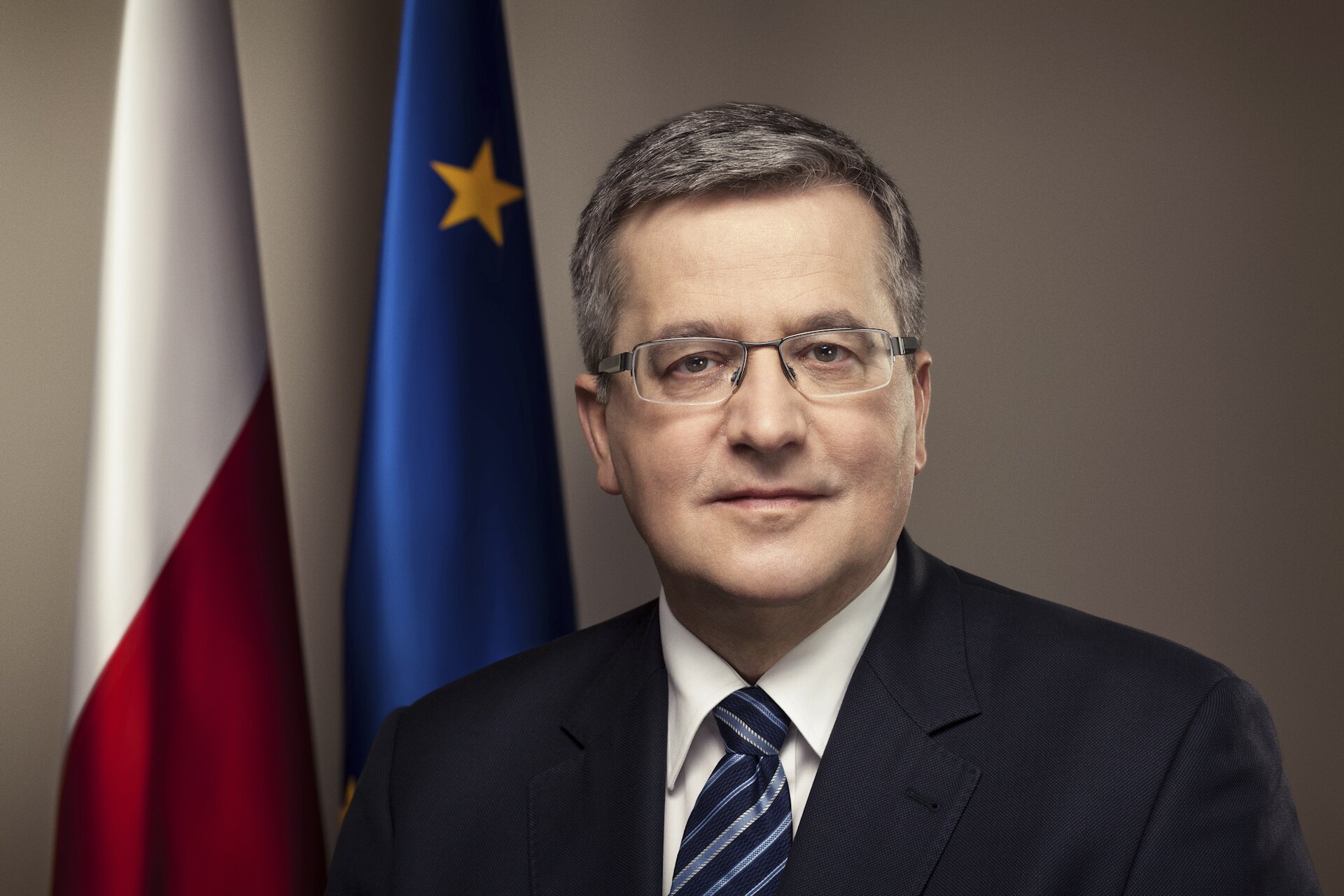 Kliknij żeby powiększyć Na zdjęciu znajduje się prezydent Bronisław Komorowski. W tle flagi: Polski i Unii Europejskiej. Mężczyzna z siwymi włosami i okularami, który ubrany jest w garnitur.