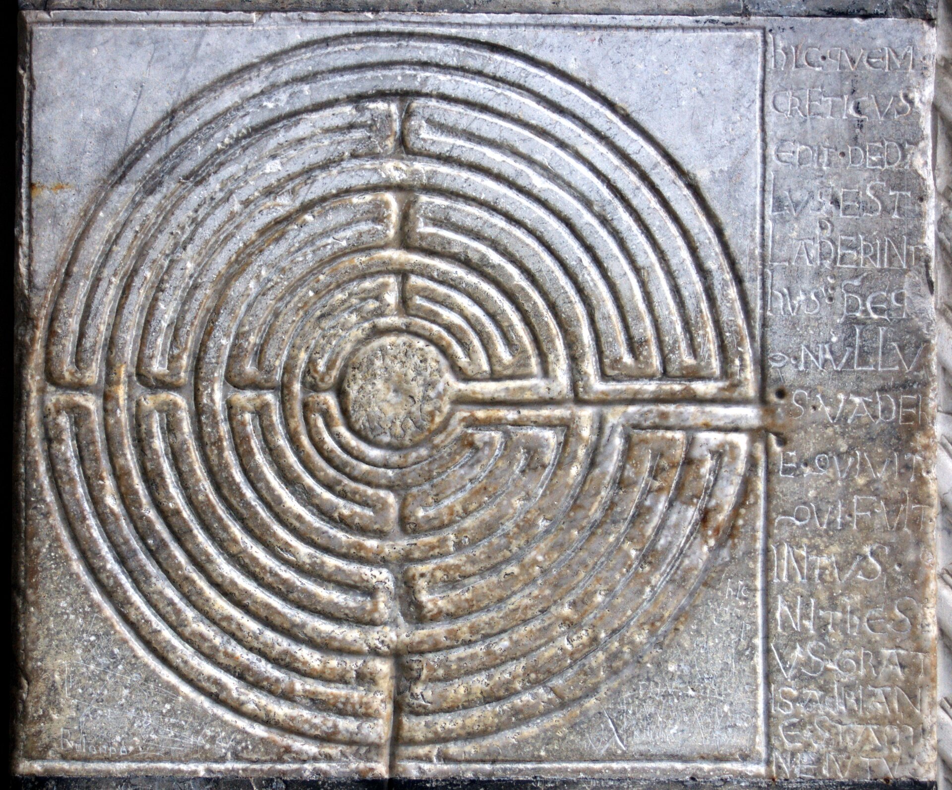 Zdjęcie przedstawia linie wyryte w kamieniu. Tworzą kształt kolistego labiryntu. Z prawej strony widoczne są zatarte napisy w obcym języku.