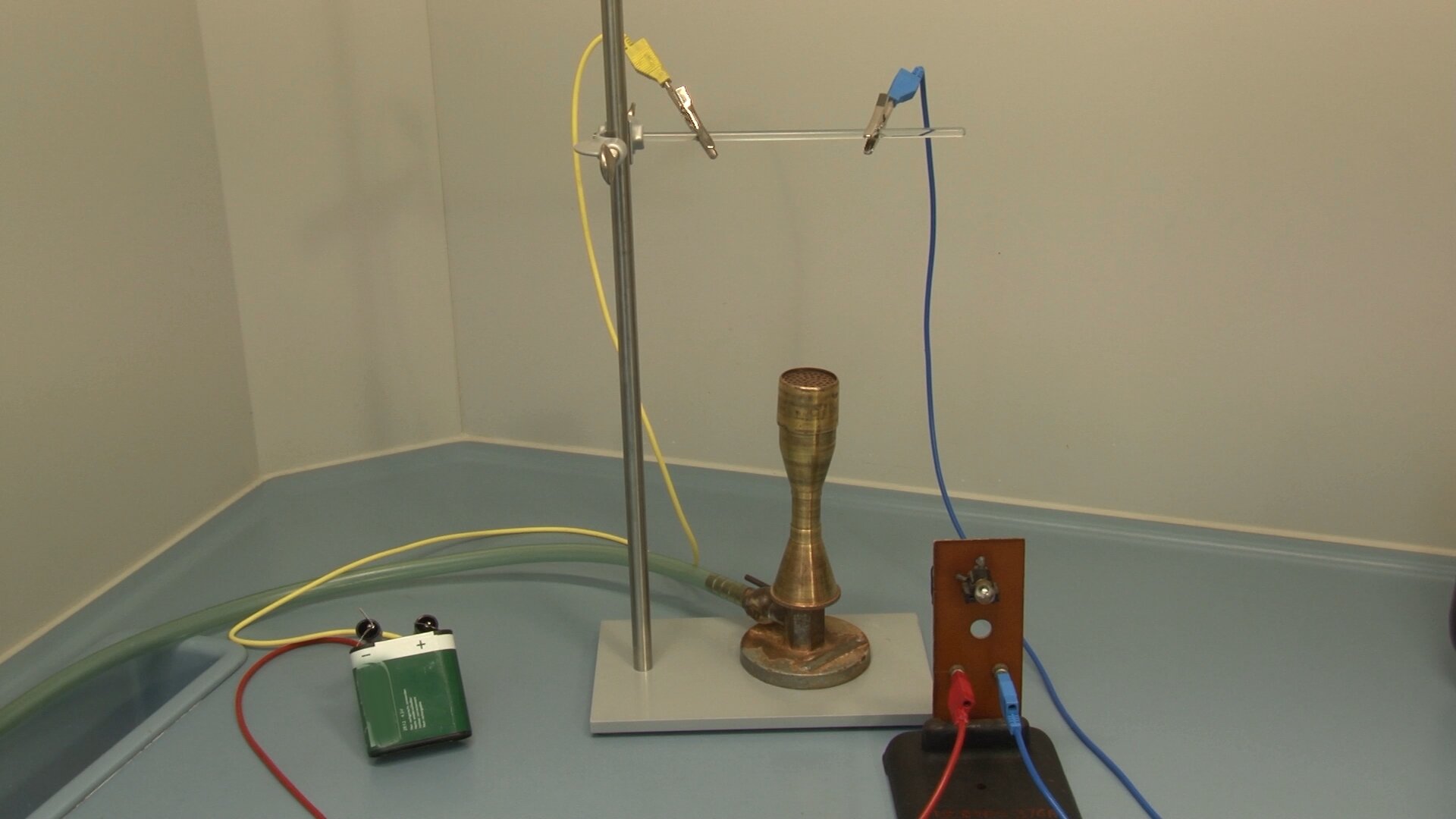 Zdjęcie prostego obwodu elektrycznego. Po lewej stronie zielona, szeroka bateria. Od jej bieguna dodatniego poprowadzono żółty przewód zakończony krokodylkiem. Krokodylek zaciśnięty jest na rurce umocowanej na statywie. Do rurki też przymocowany jest też krokodylkiem niebieski przewód. Przewód ten połączony jest końcówką typu bananek z  żarówką przymocowaną do stojaka. Od tej żarówki odchodzi, również podłączony banankiem, czerwony przewód. Czerwony przewód poprowadzony jest do ujemnego bieguna baterii.