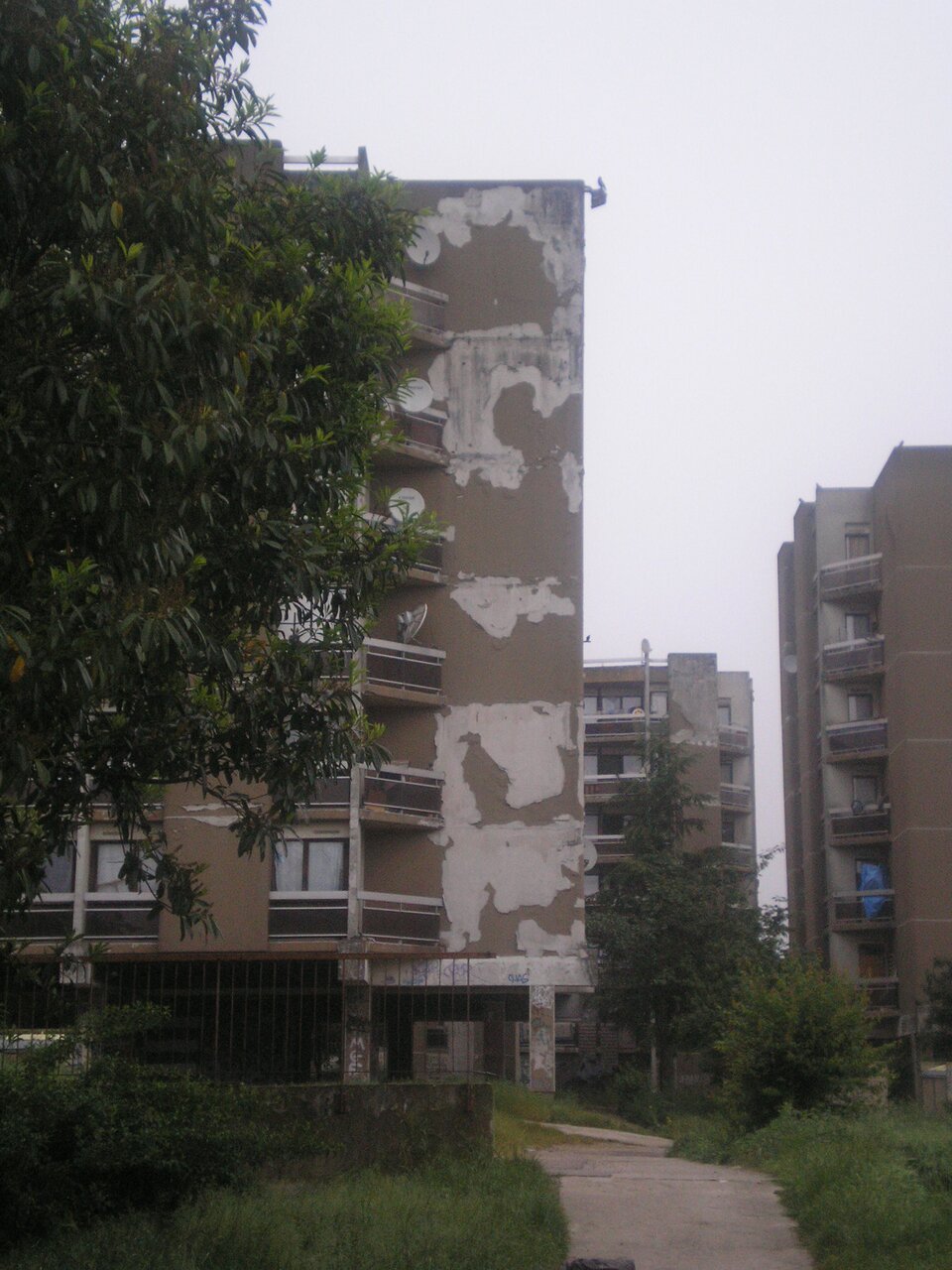 Zdjęcie przedstawia blokowisko we Francji. Jest wysoki, z jego ścian odchodzi farba i tynk. Ogrodzenie jest stare.