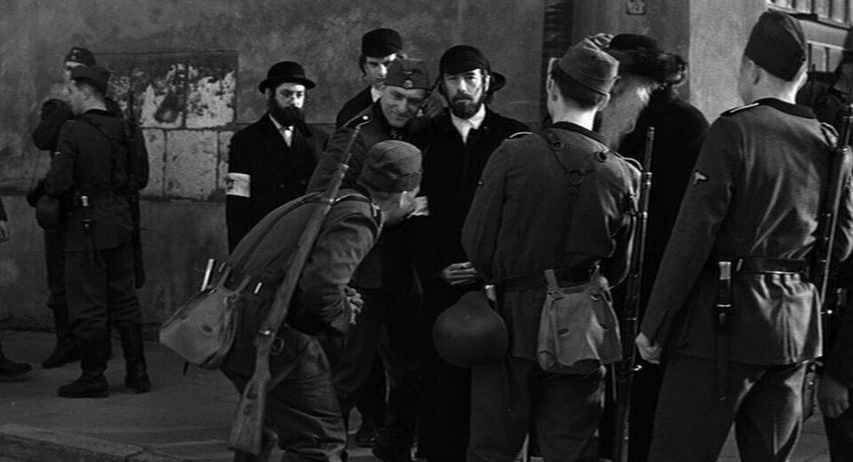 Zdjęcie przedstawia ulicę. Stoi na niej grupa Żydów w tradycyjnych strojach. Są otoczeni przez niemieckich żołnierzy.
