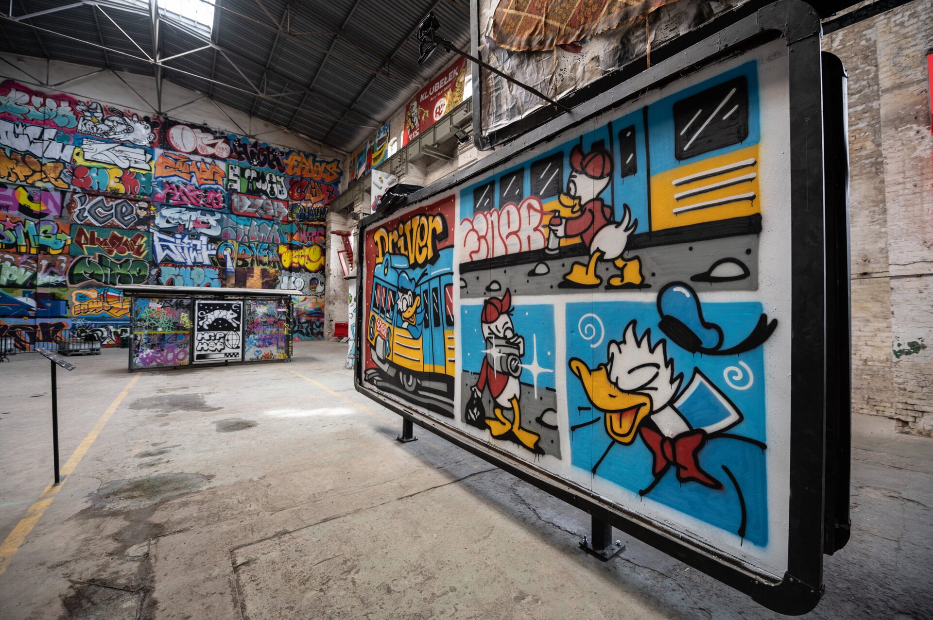 Na zdjęciu znajduje się wysokie pomieszczenie o jasnych, surowych ścianach i podłodze. Na pierwszym planie jest billboard [czytaj: bilbord], który ustawiony jest bokiem. Na billboardzie [czytaj: bilbordzie] namalowane jest komiksowe graffiti [czytaj: grafiti] z rysunkami kaczek składające się z 4 części. Na pierwszej części jest fragment niebiesko‑żółtego pociągu, w którym jest Kaczor Donald w niebieskiej czapce. Nad pociągiem jest żółty napis „Driver [czytaj: drajwer]”. Tło jest w kolorze czerwonym. Na drugiej części jest biała kaczka z żółtym dziobem i łapami, która ubrana jest w czerwoną bluzkę i czapkę. W lewej dłoni trzyma czerwony spray, którym maluje po niebiesko‑żółtym wagonie. Na trzeciej części jest biała kaczka z żółtym dziobem i łapami, która ubrana jest w czerwoną bluzkę i czapkę. W lewej dłoni trzyma szary aparat, a w prawej czarną torbę. Tło jest w kolorze niebiesko‑szarym. Na czwartej części jest popiersie Kaczora Donalda na niebieskim tle. Na drugim planie fotografii jest przystanek tramwajowy widziany od tyłu (ten, którego przód widać na poprzednim zdjęciu), na którym jest kilka kolorowych graffiti [czytaj: grafiti]. W tle na ścianach jest wiele graffiti [czytaj: grafiti]. 