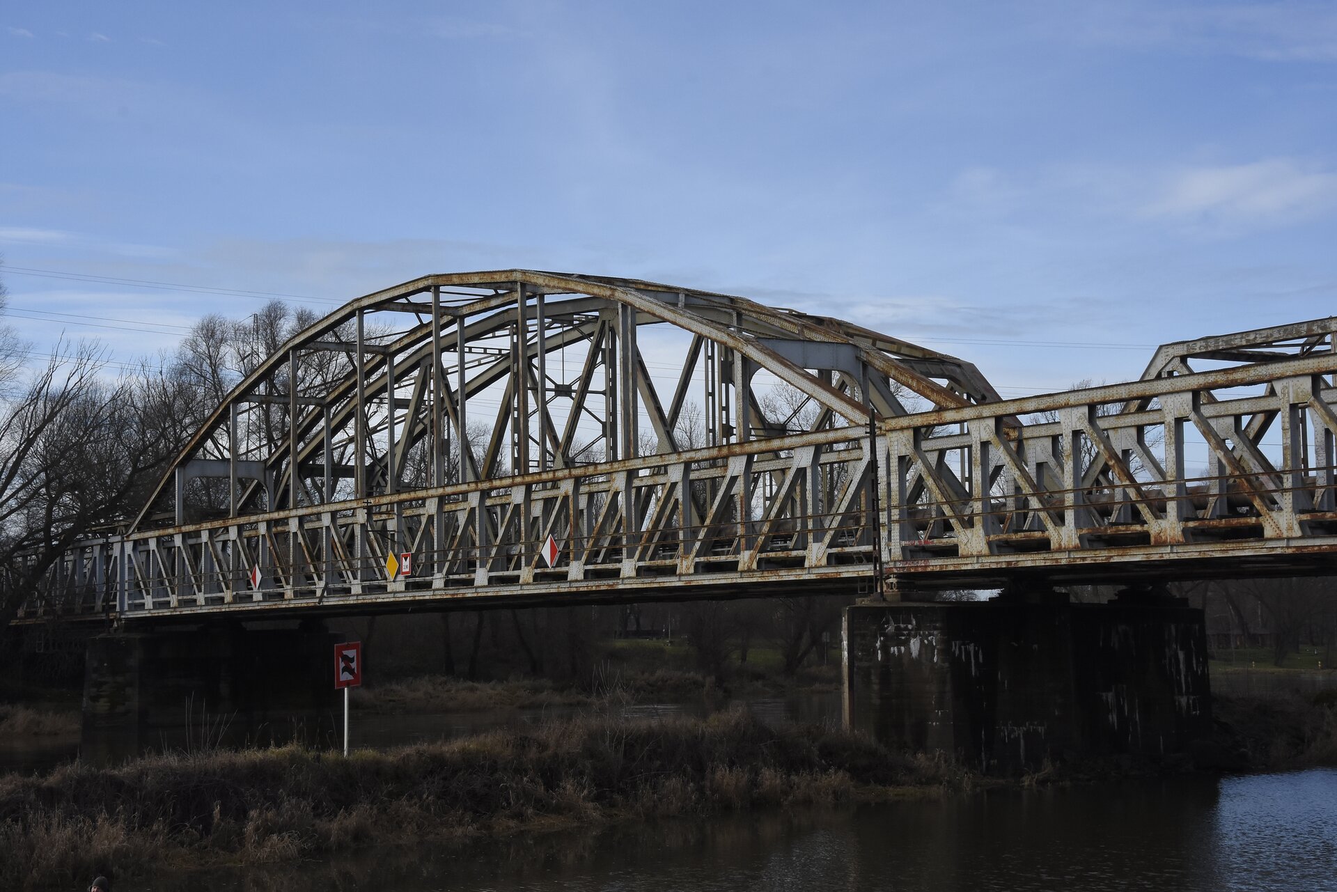 Most stalowy łukowy z jazdą dołem. Widoczny jest fragment mostu z jednym z łuków. Stalowy łuk podtrzymuje od góry platformę z torami. Platforma rozciągnięta jest nad rzeką między dwoma betonowymi filarami. W tle widoczne są drzewa po dalszej stronie mostu.