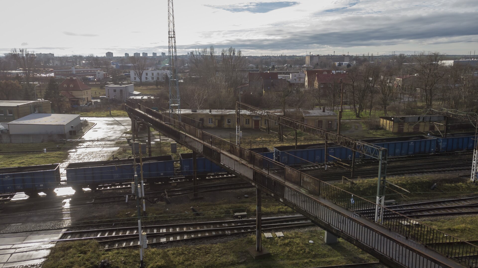  Ilustracja przedstawia widok z boku na kładkę dla pieszych, która znajduje się nad torowiskiem składającym się z dziesięciu torów. Kładka jest wykonana z metalu, a do niej prowadzą schody. Na niektórych elementach kładki widoczna jest rdza. Szlabany na przejeździe kolejowym są zamknięte. Na jednym z torów przejeżdża pociąg towarowy, widocznych jest sześć wagonów towarowych znajdujących się pod kładką. Na torowisku widać także semafory. W tle widoczne są wysoki przekaźnik, miasto oraz chmury. Kładka umożliwia pieszym bezpieczne przekroczenie torów kolejowych.