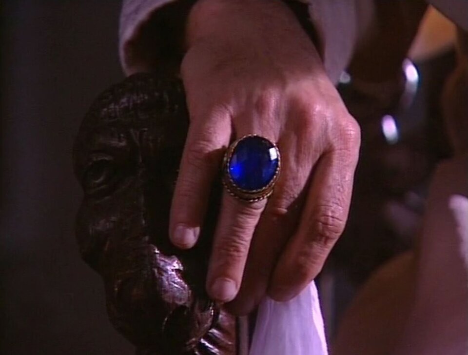Zdjęcie przedstawia kadr z filmu. To zbliżenie na prawą, męską dłoń spoczywającą na rzeźbionym oparciu fotela. Na serdecznym palcu znajduje się duży, złoty pierścień z szafirem. Dłoń trzyma jeszcze białą, materiałową chusteczkę.