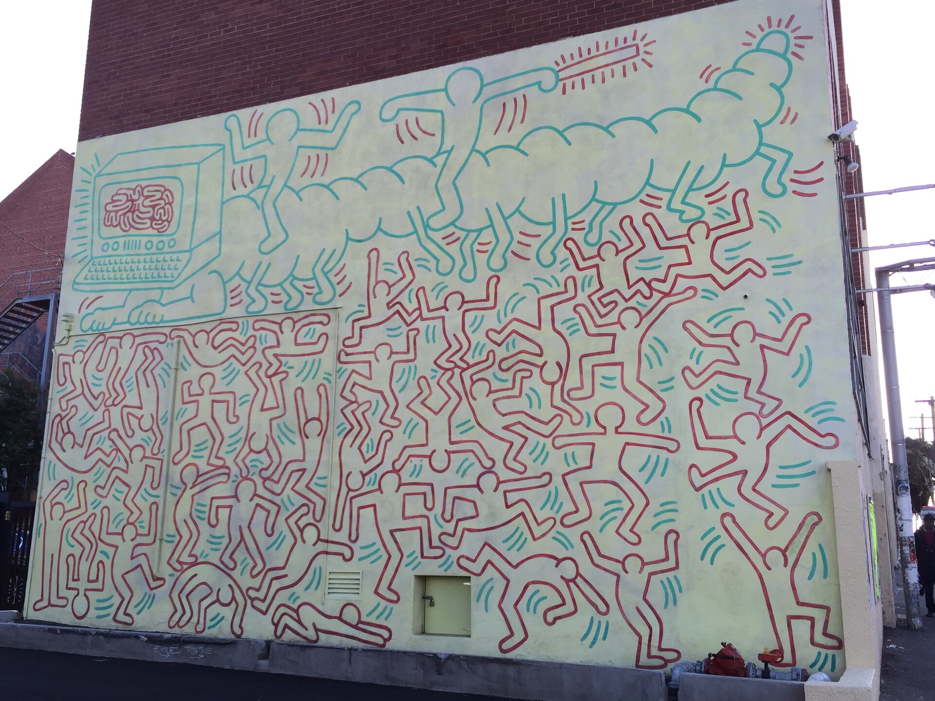 Fotografia przedstawia mural na ścianie budynku autorstwa Keith Haring. Ściana w białym kolorze pokryta jest płaskimi postaciami o czerwonych konturach, niewypełnionymi kolorem, są to schematyczne sylwetki. Postacie przybierają różne pozy, przeważnie jest to wyskok lub podskok. Nad nimi idzie gąsienica o niebieskich konturach, z komputerem zamiast głowy, na ekranie komputera przedstawiony jest czerwony mózg. Na grzbiecie gąsienicy jadą dwie postacie, jedna z nich trzyma podłużny przedmiot od którego odchodzą kreski przypominające promyki. Mural znajduje się na bocznej ścianie budynku z czerwonej cegły, w dolnej części ściany są małe beżowe, drzwiczki.
