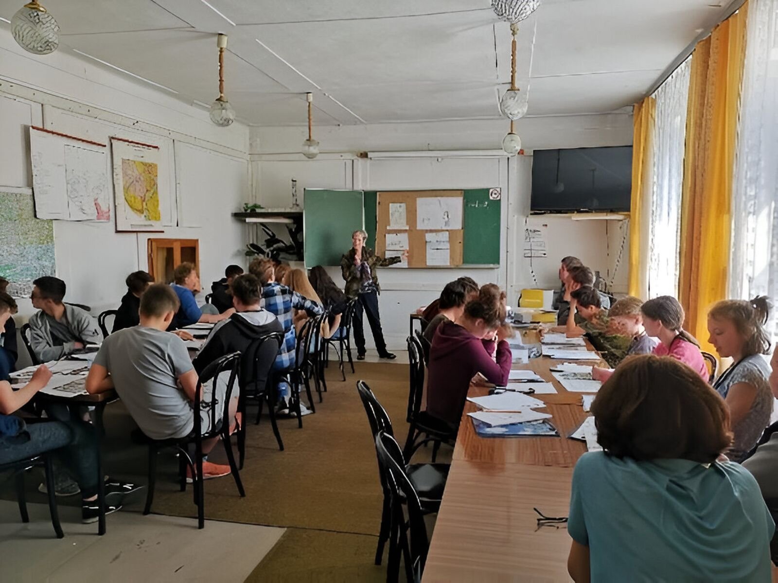 Zdjęcie przedstawia klasę. Po lewej i prawej stronie ułożone równolegle ławki, przy nich uczniowie robiący notatki. W oddali na ścianie zielona tablica, na niej poster składający się z czterech kartek, przy nim nauczyciel wskazujący na tablicę.