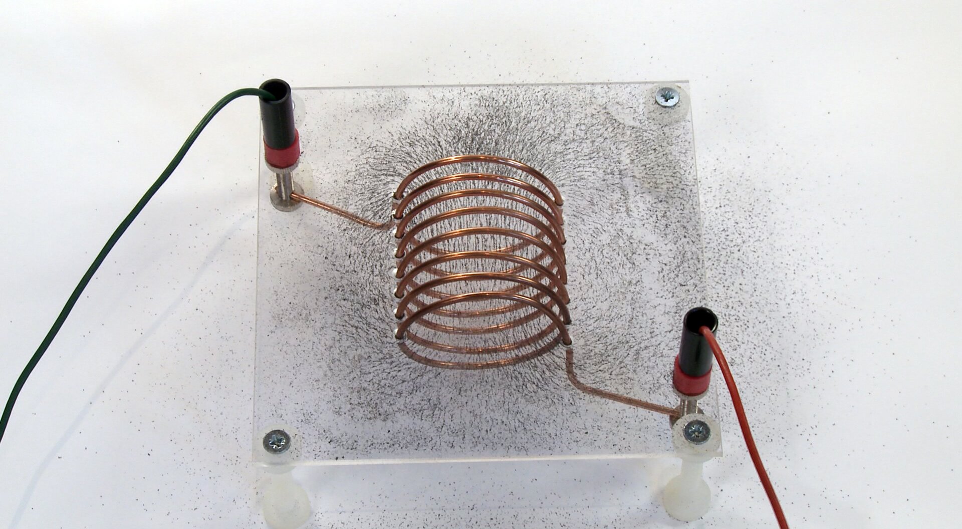 Rys. 3. Na zdjęciu widać miedzianą zwojnicę w postaci spirali, umieszczoną na plastikowym, przezroczystym stoliczku. Zwojnica podłączona jest do prądu dwoma przewodami. Zwojnica zamontowana jest na stoliku w taki sposób, że stolik przechodzi przez jej środek wzdłuż długości zwojnicy. Na stoliku umieszczone są opiłki żelaza. Zwojnica z prądem stanowi źródło pola magnetycznego. Opiłki żelaza oddziałujące z polem magnetycznym ustawiają się w taki sposób, że wewnątrz zwojnicy tworzą linie równoległe do długości spirali a na zewnątrz są rozbieżne i obiegają zwojnicą dookoła. 