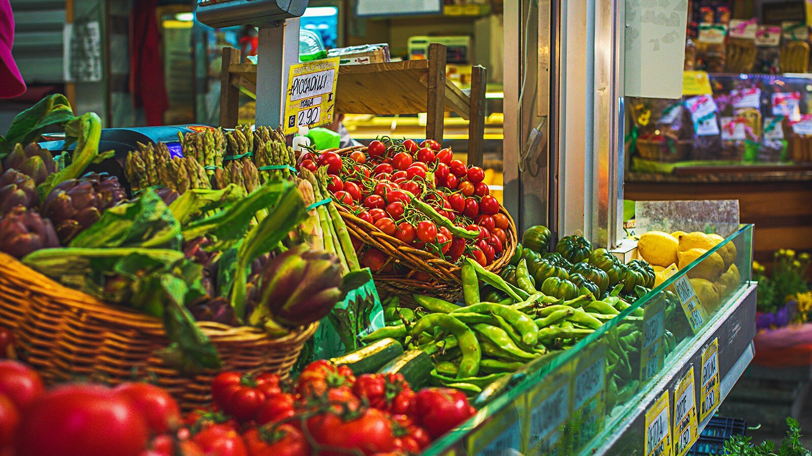 Zdjęcie przedstawia wnętrze targowiska. Po lewej stronie i w środku widoczna jest długa lada, na której wystawione są różne warzywa, ułożone luzem lub w wiklinowych koszach. Widoczne są czerwone pomidory, zielone strąki fasoli, zielone pomidory, cytryny, małe pomidorki koktajlowe, pęczki zielonych szparagów, karczochy. Na froncie lady widoczne są żółte karteczki z białymi napisami informującymi skąd pochodzi dany produkt oraz jaka jest jego cena. W tle widoczne są inne stoiska targowiska.