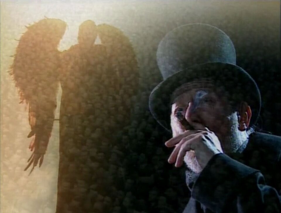 Zdjęcie przedstawia kadr z filmu. Na pierwszym planie znajduje się palący cygaro starszy mężczyzna. Jest ubrany w czarny płaszcz, na głowie ma czarny cylinder. Za jego plecami, na ścianie znajduje się rozświetlony cień dużego, skrzydlatego anioła.