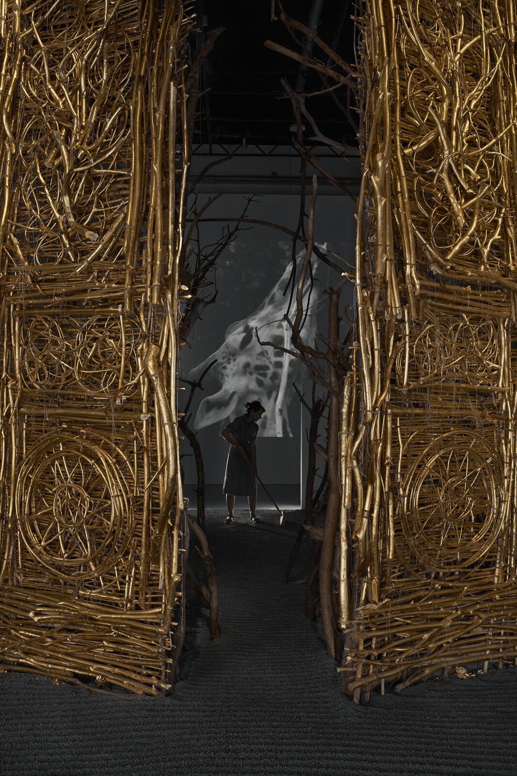 Na zdjęciu znajduje się praca Agnieszki Kalinowskiej, która ustawiona jest na szarej podłodze. Przedstawia ozdobne, złote, dwuskrzydłowe drzwi wykonane z gałązek. Drzwi są uchylone. Na drugim planie znajduje się kobieta w sukience za kolano. W dłoniach trzyma miotłę. Obok niej są pnie drzew. W tle znajduje się duży czarno‑biały obraz przedstawiający głowę żyrafy. 