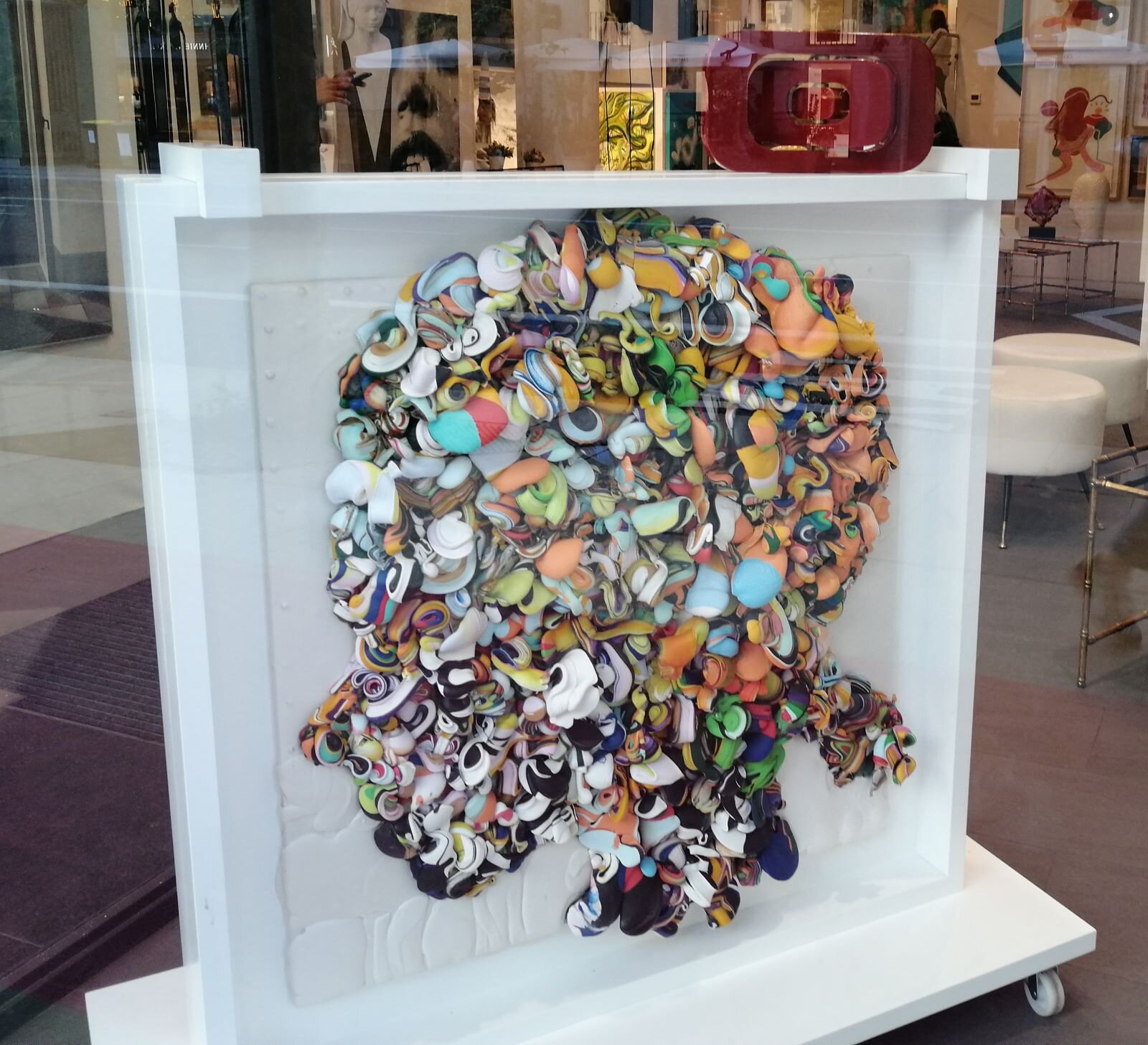 Zdjęcie przedstawia ekspozycję na wystawie sklepowej: to oszklona witryna w kształcie kwadratu, z umieszczoną w środku formą przestrzenną. Jest to głowa dziewczyny z profilu, zrobiona z umieszczonych blisko siebie różnych kolorowych, niewielkich przedmiotów, które trudno rozpoznać, bo są pozginane. Jest to nowoczesna płaskorzeźba z materiałów odzyskanych, czyli recyklingowa.