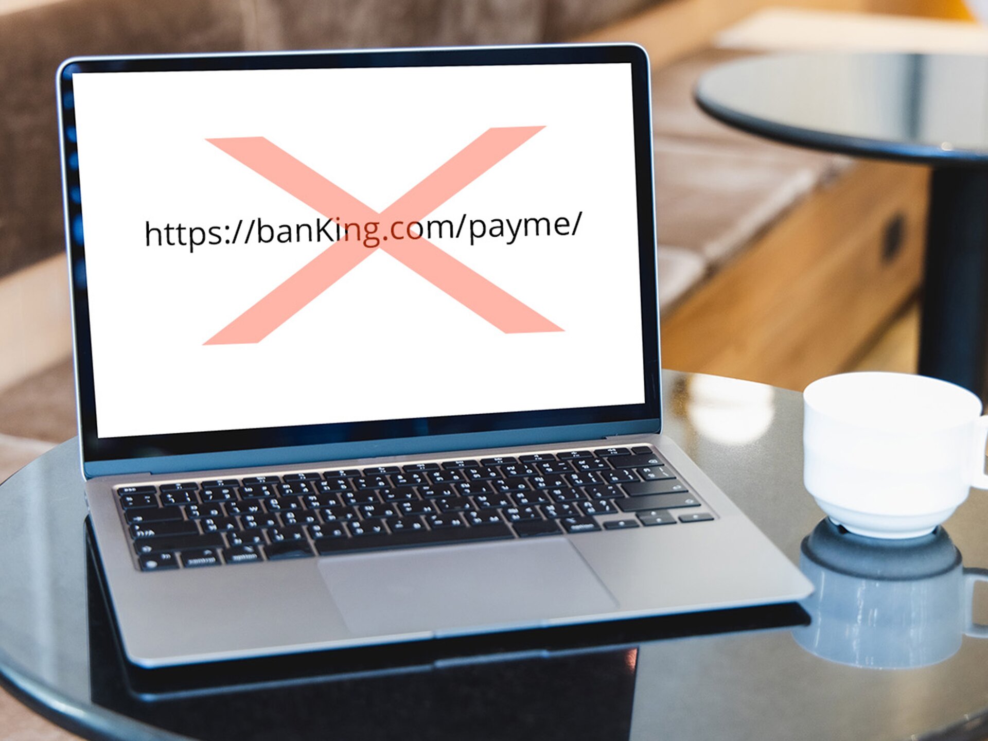 Zdjęcie przedstawia otwartego laptopa, który stoi na blacie okrągłego stolika. Na ekranie laptopa widoczny jest adres strony internetowej: https://banKing.com/payme/ przekreślony dużym, czerwonym znakiem X. Na prawo od laptopa widoczna jest biała filiżanka. W tle widoczny fragment kolejnego okrągłego stolika oraz fragment ławy do siedzenia.