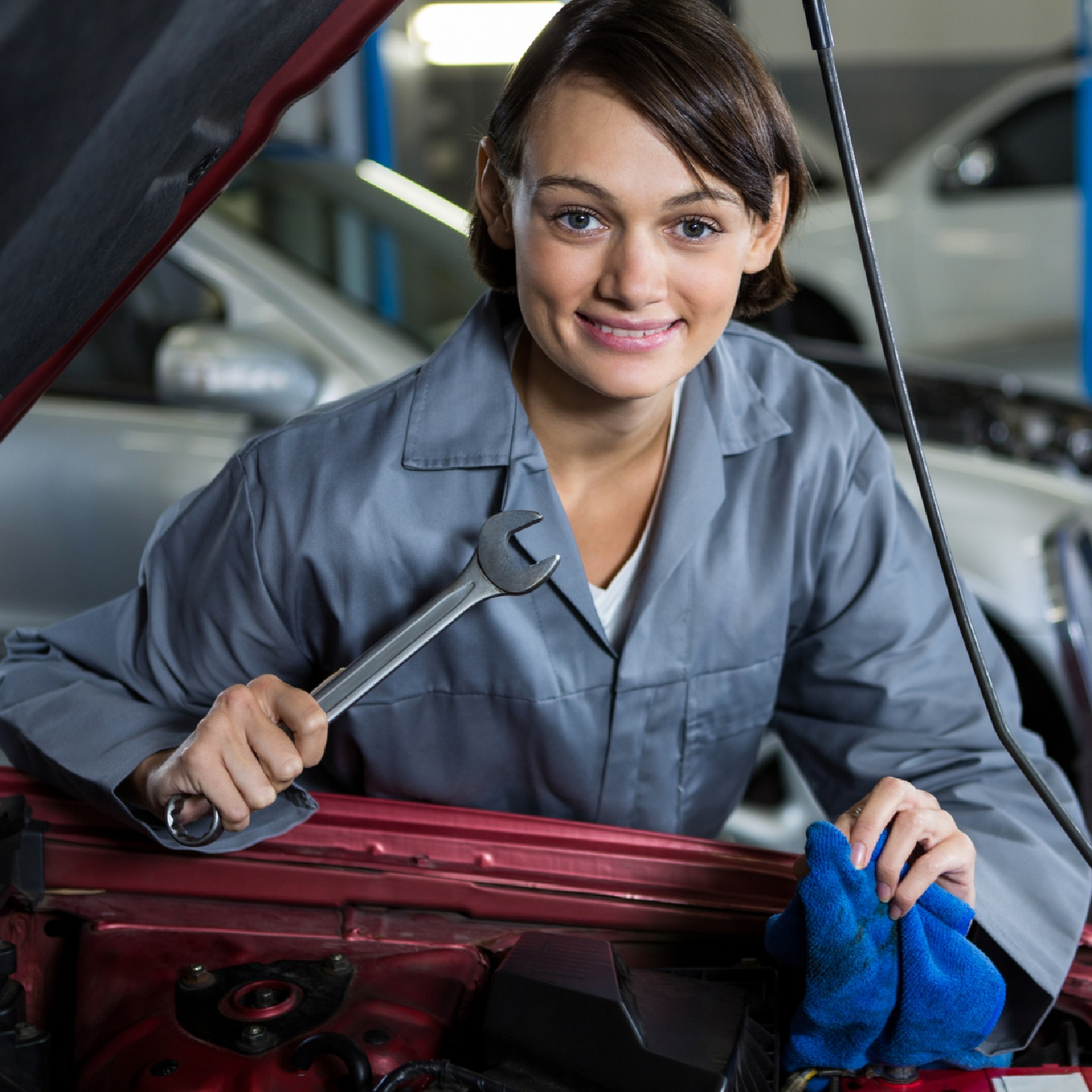 Ilustracja przedstawia panią mechanik, ubraną w niebieski kombinezon roboczy. Pochyla się nad otwartą maską samochodu, trzymając w dłoni klucz naprawczy. W drugiej dłoni, trzyma niebieską szmatkę. W tle widoczne są inne samochody , z otwartymi maskami.