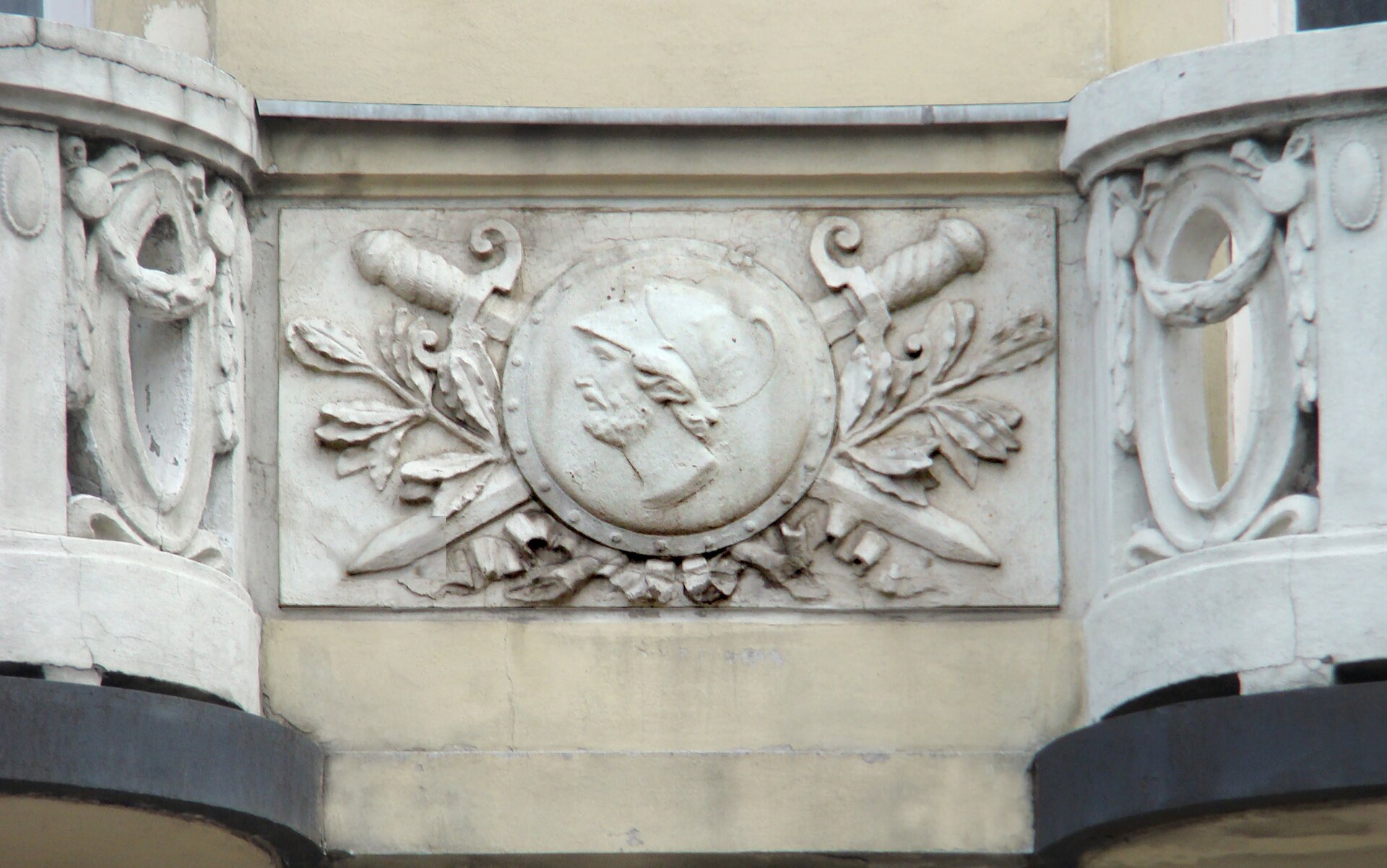 Ilustracja przedstawia fragment budynku, na którym umieszczono elementy dekoracyjne. Na prostokątnym polu znajduje się w centralnej części okrągła tarcza z wyrytą głową żołnierza z lewego profilu. Za tarczą umieszczono dwa krzyżujące się miecze, z rękojeściami w górnej części. Pomiędzy tymi elementami umieszczono jeszcze liście na długich gałązkach. Z obu stron zdjęcia znajdują się balkony z dekoracyjnymi barierkami.