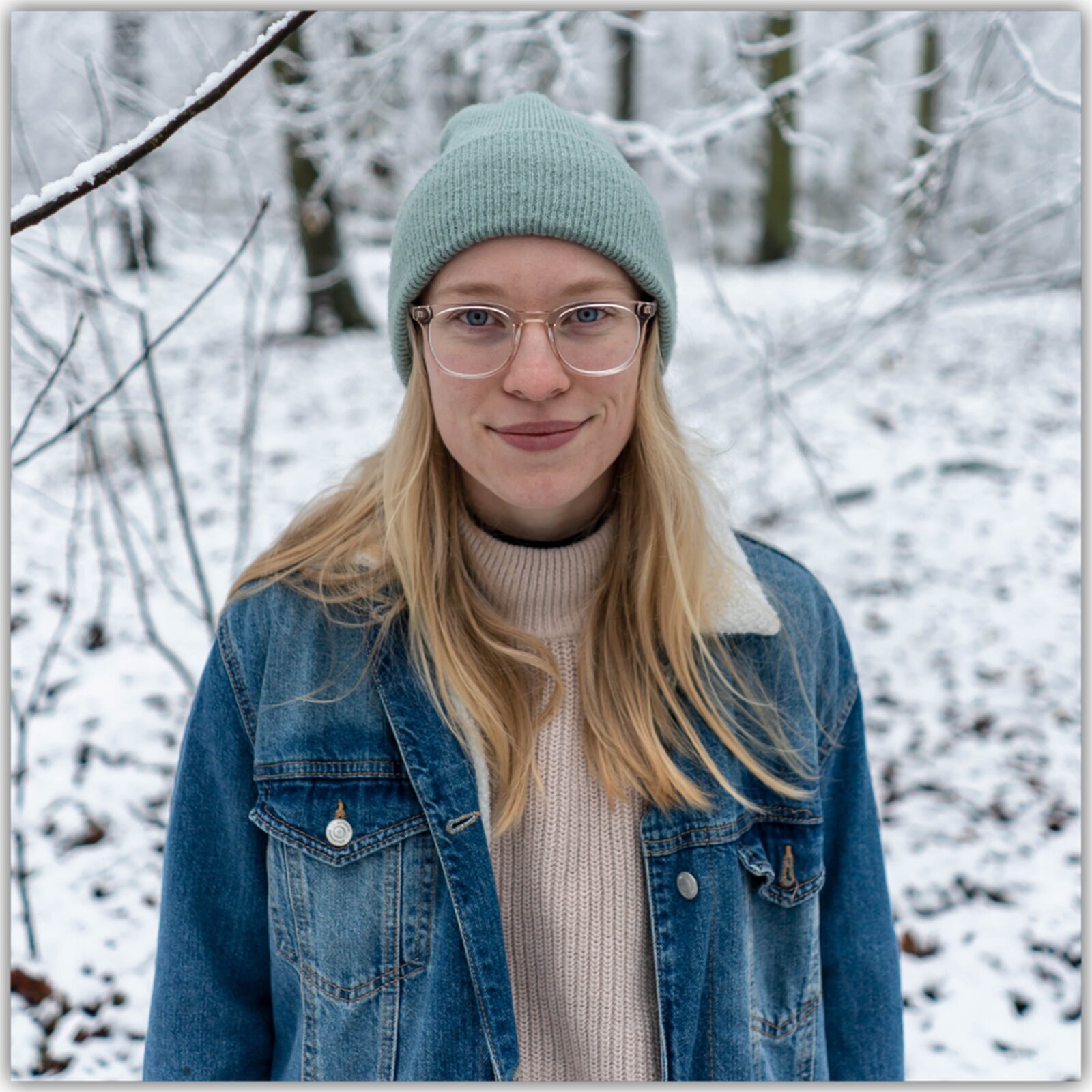 Grafika przedstawia młodą kobietę w długi, prostych blond włosach i okularach. Ma na sobie jasno zieloną czapkę, kurtkę dżinsową a pod nią beżowy sweter z golfem. Stoi na tle ośnieżonego lasu. Uśmiecha się.
