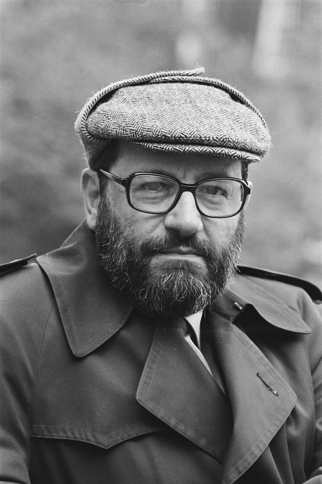 Czarno-białe zdjęcie przedstawiające Umberto Eco. Ma na sobie prochowiec i kaszkiet. Nosi okulary i brodę.