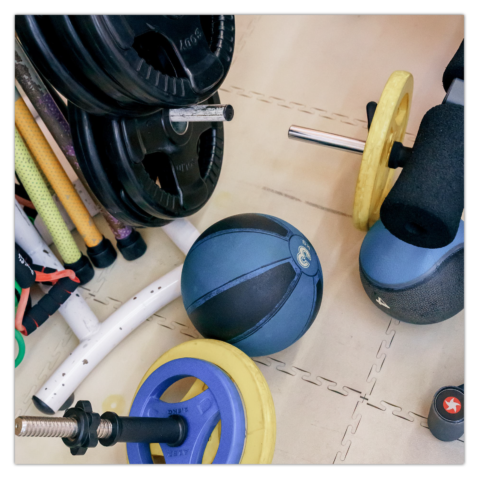 Zdjęcie przedstawia fragment  siłowni. Na podłodze leżą hantle oraz 3 kilogramowa piłka. Na stojakach, ustawione są ciężarki o różnej wadze.