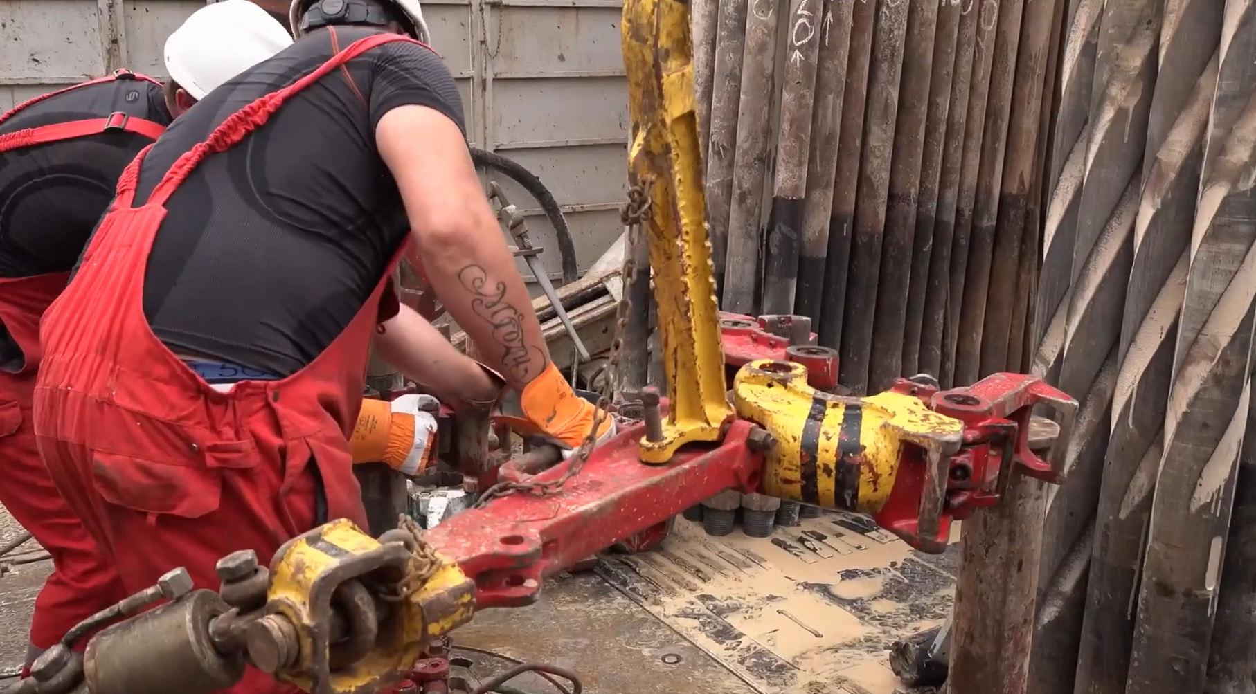 Ilustracja interaktywna. Ilustracja przedstawia masywny, czerwony klucz maszynowy do przewodu wiertniczego. Klucz maszynowy jest w kształcie szczypiec. Klucz podwieszony jest na linie w szybie wiertniczym, z wykorzystaniem przeciwwag. Z jego pomocą stołowi skręcają i rozkręcają połączenia gwintowe przewodu wiertniczego. Klucz maszynowy składa się z: wieszaka klucza maszynowego służącego do podwieszenia klucza na łańcuchu w szybie wiertniczym, szczęki klucza maszynowego która jest elementem zapinanym na zworniku skręcanego elementu rurowego oraz ostrza klucza maszynowego który jest elementem odpowiedzialnym za zwiększenie tarcia między szczękami a zwornikiem skręcanego elementu.